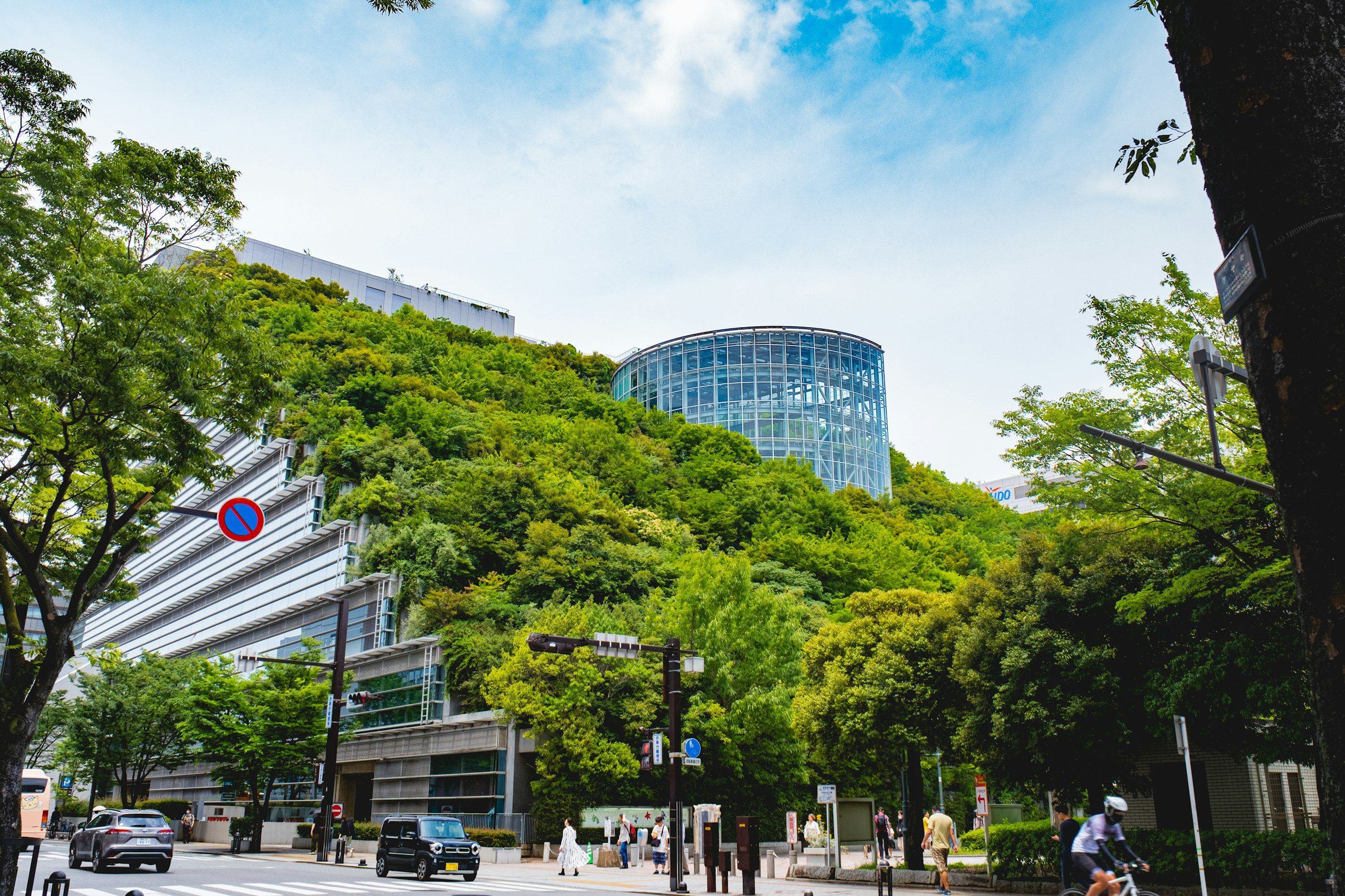 Urban scene with buildings, trees, and a blue sky. People walking on the sidewalk, cars parked on the street, and traffic signals visible.
