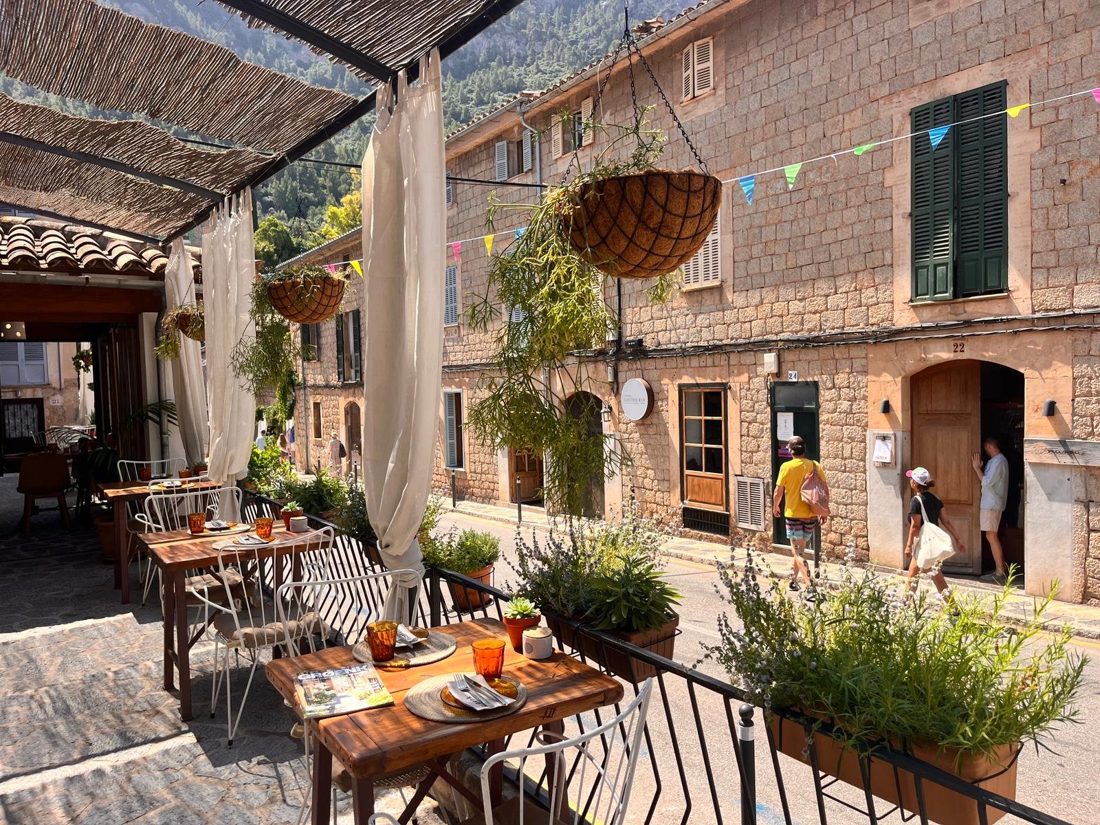 An outdoor restaurant patio with wooden tables set with dishes and glasses, surrounded by potted plants. Hanging baskets with plants and colorful pennant banners are visible, along with a stone building and people walking by.