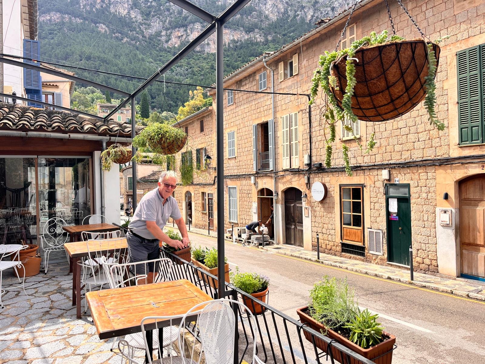 Man standing on a patio with outdoor furniture, potted plants, and hanging flower baskets, overlooking a street with old brick buildings and mountain scenery in the background.