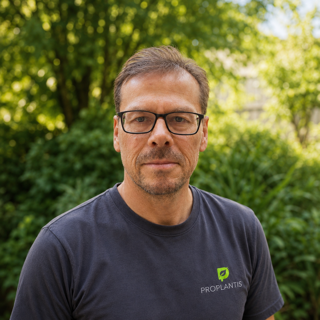 A middle-aged man with glasses and short brown hair standing outdoors in front of greenery, wearing a black PROPLANTIS t-shirt.