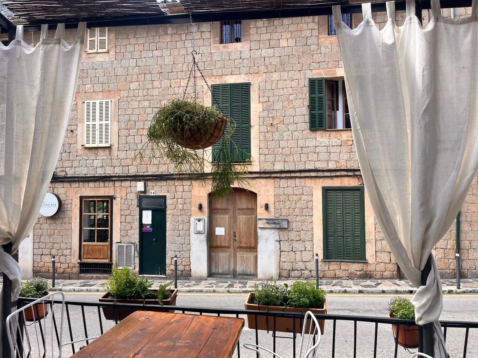 View from a patio looking out onto a stone building with green shutters, a hanging potted plant, and a wooden door, framed by white curtains.