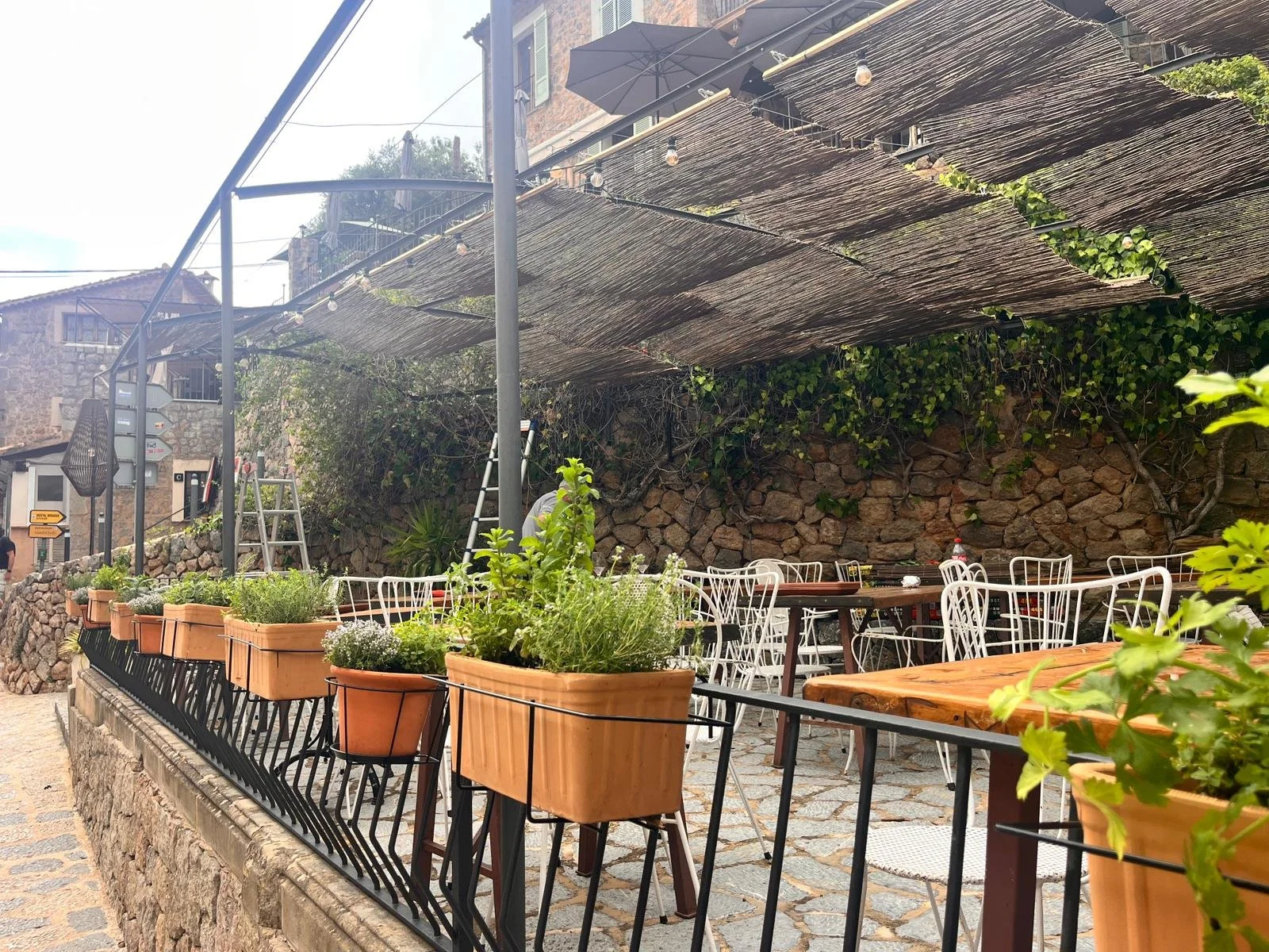 Outdoor patio with potted plants, metal chairs, wooden tables, and a shaded bamboo roof, surrounded by stone walls and greenery.