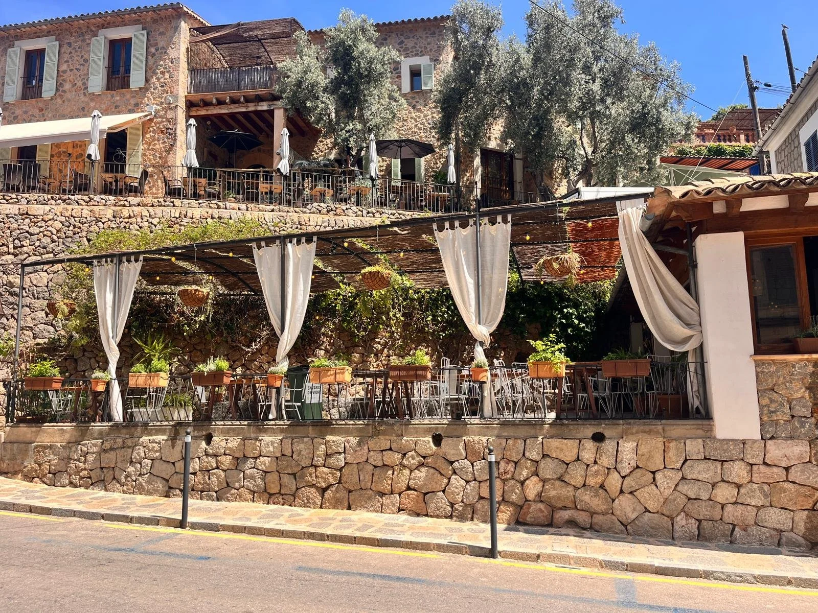 Outdoor dining area with white curtains, potted plants, and string lights, set against a stone building with balconies and trees in a sunny setting.