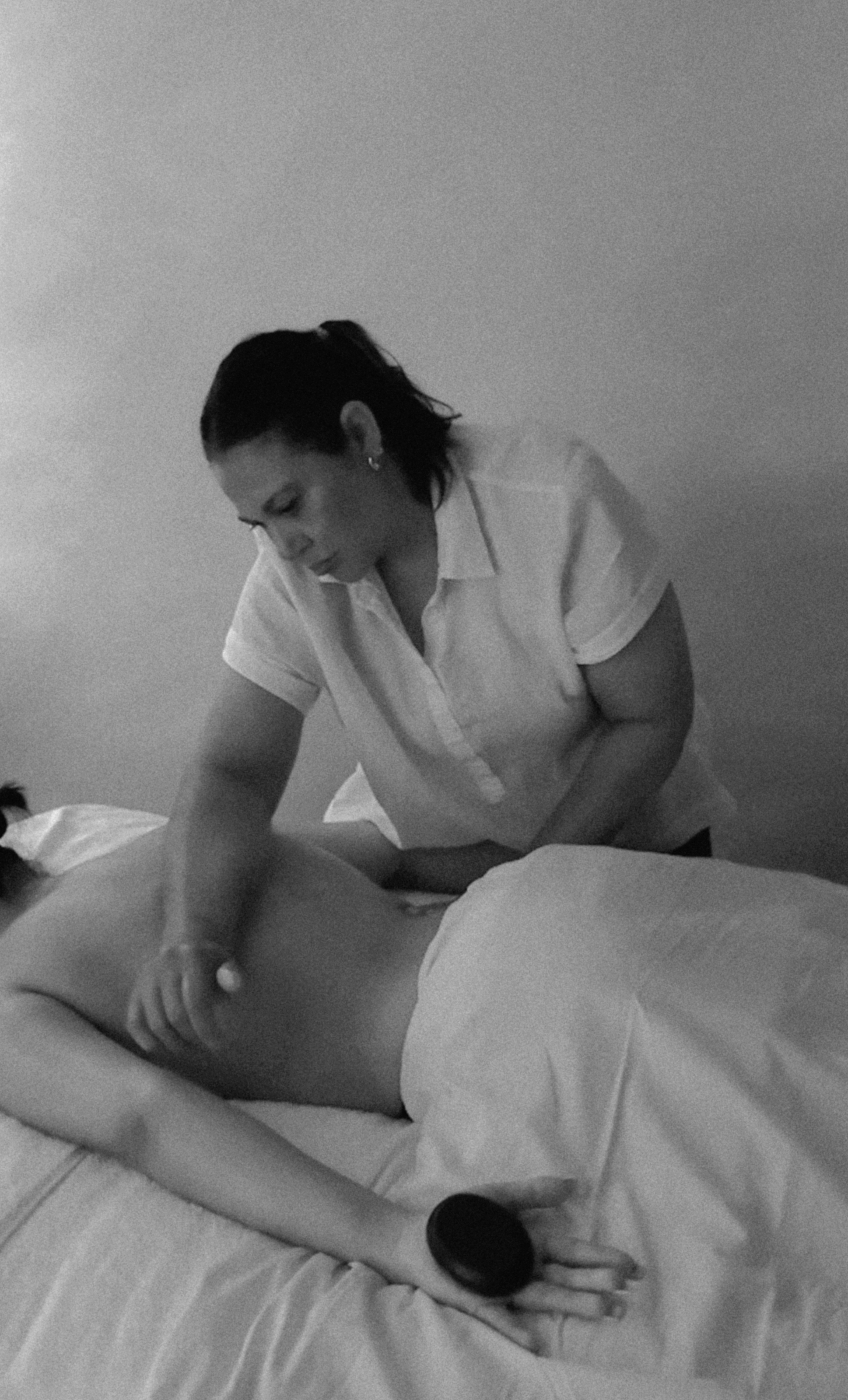 A woman in a white shirt gives a massage to a person lying face down on a bed, in black and white.