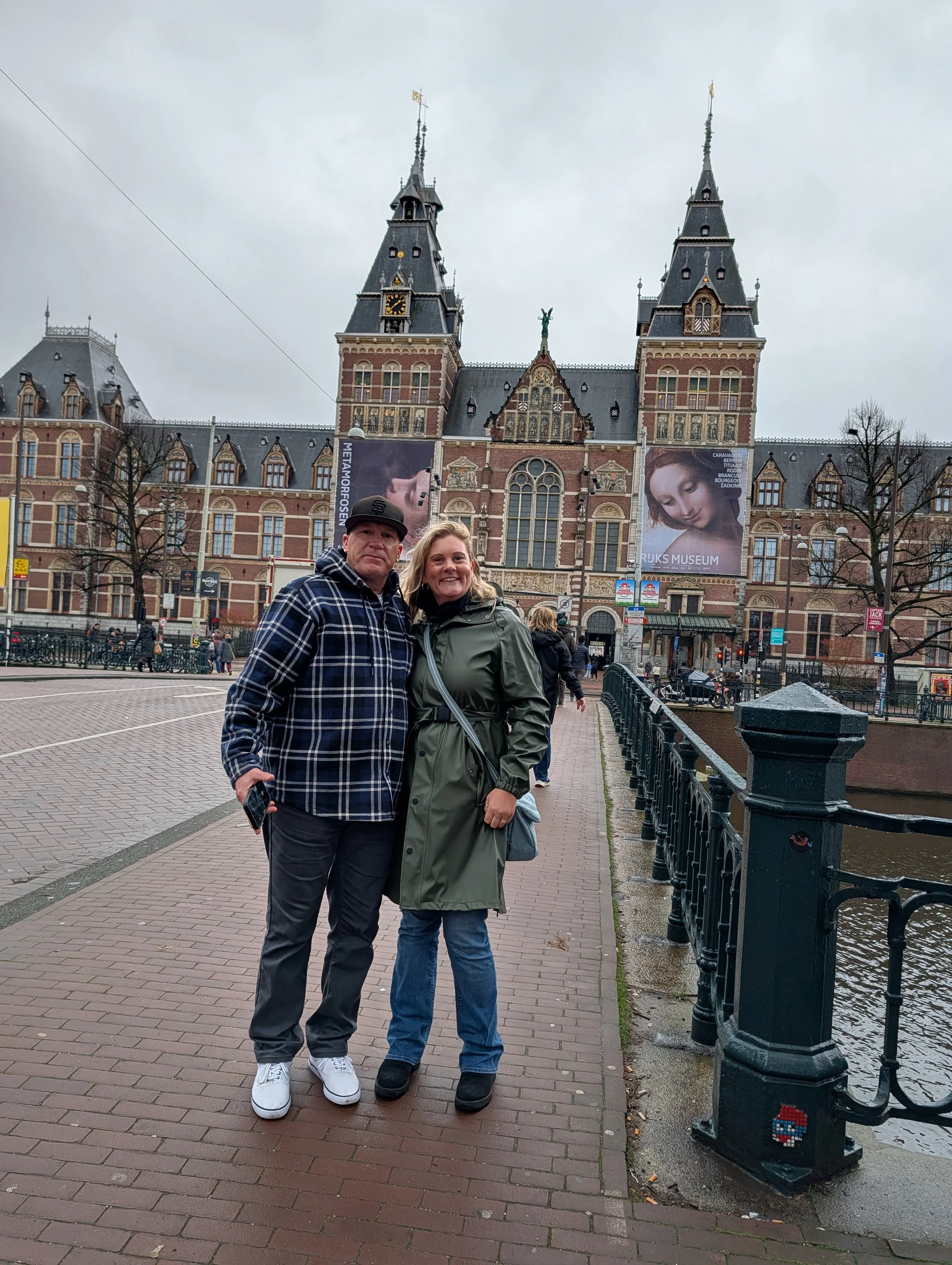 Two people standing on a brick sidewalk in front of a historic brick building with two towers, a clock, and a statue on its roof. The sky is cloudy, and there are other pedestrians in the background.