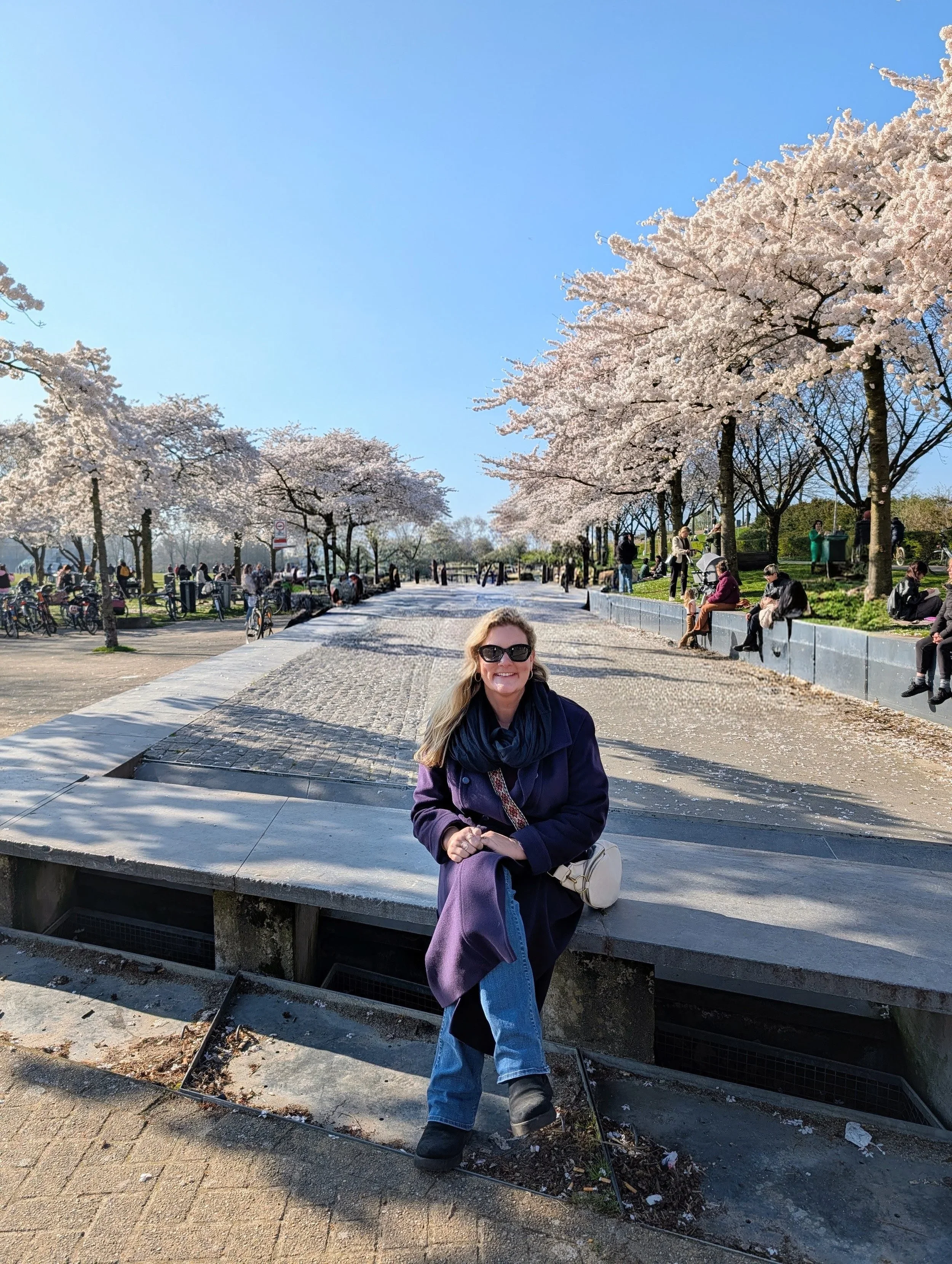 A woman with long hair, wearing sunglasses and dark clothing, smiling and standing in front of blooming cherry blossom trees in a park on a sunny day.