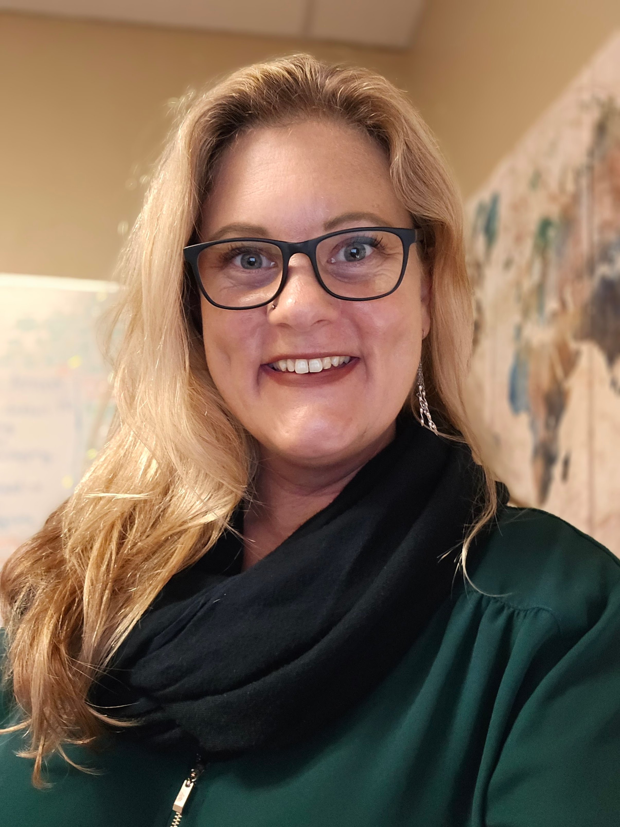 Close-up of a smiling woman with glasses, long blonde hair, wearing a black and emerald green jacket, indoors with a colorful painting in the background.