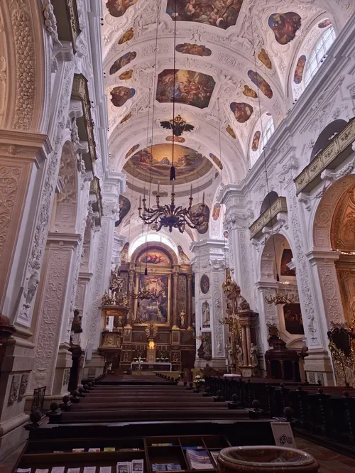 Interior of a grand church with high ornate white walls and ceiling adorned with painted religious scenes, a large chandelier hanging from the ceiling, and an elaborate altar at the front.