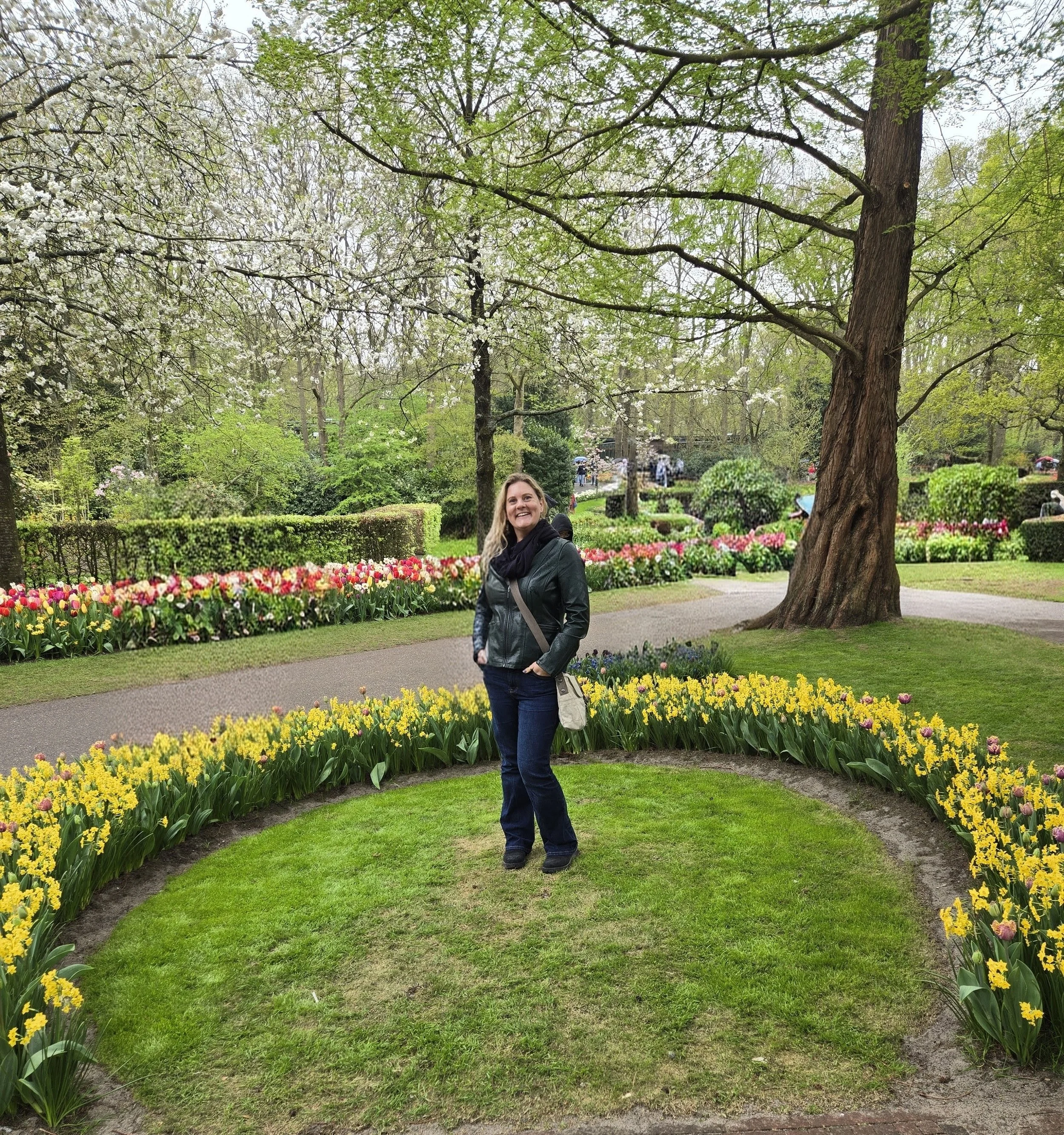 A woman smiling in a garden with colorful tulips, blooming trees, and lush greenery.