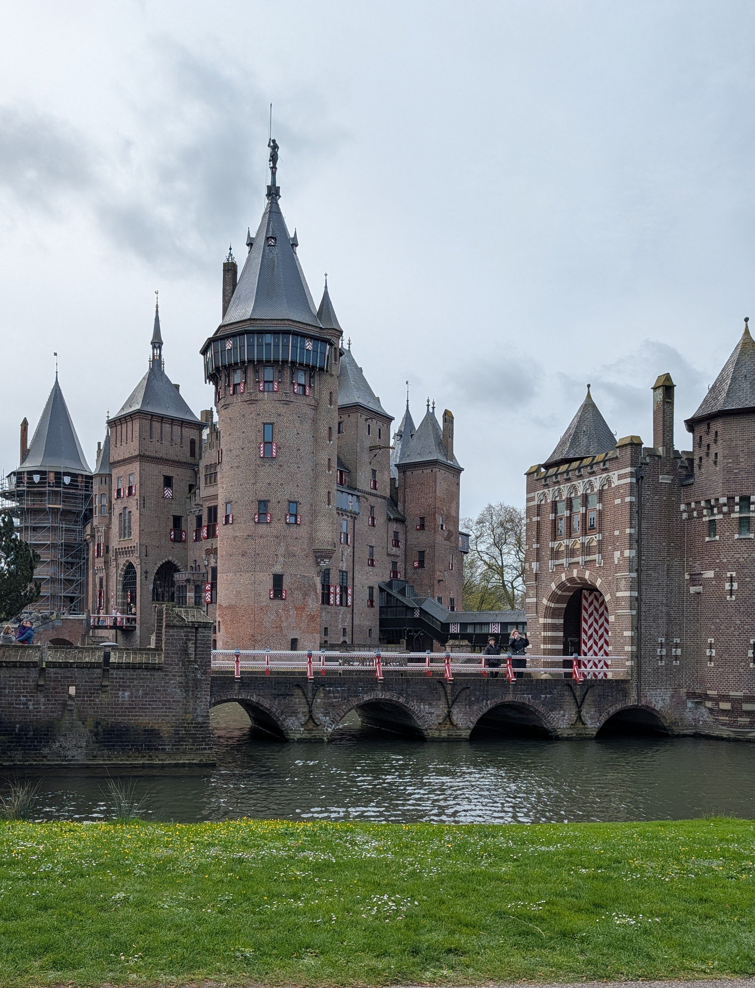 A historic castle with pointed towers and turrets, situated along a river, with a bridge in the foreground and a cloudy sky overhead.