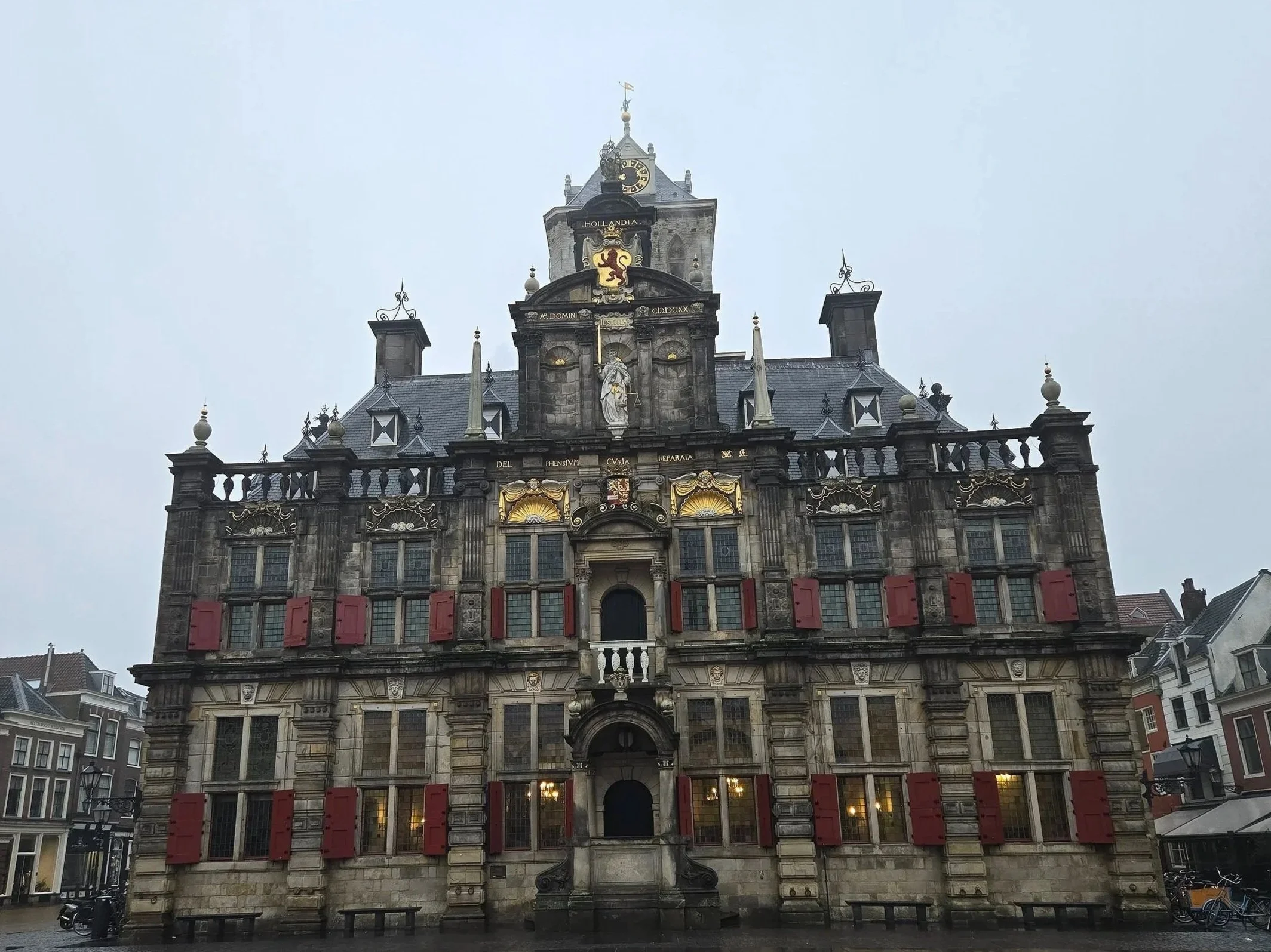 A historic stone building with elaborate architectural details, red shutters, a clock tower, and a lion crest at the top.