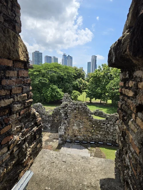 View through an opening in ancient stone ruins, showing modern skyscrapers and green trees against a partly cloudy sky.