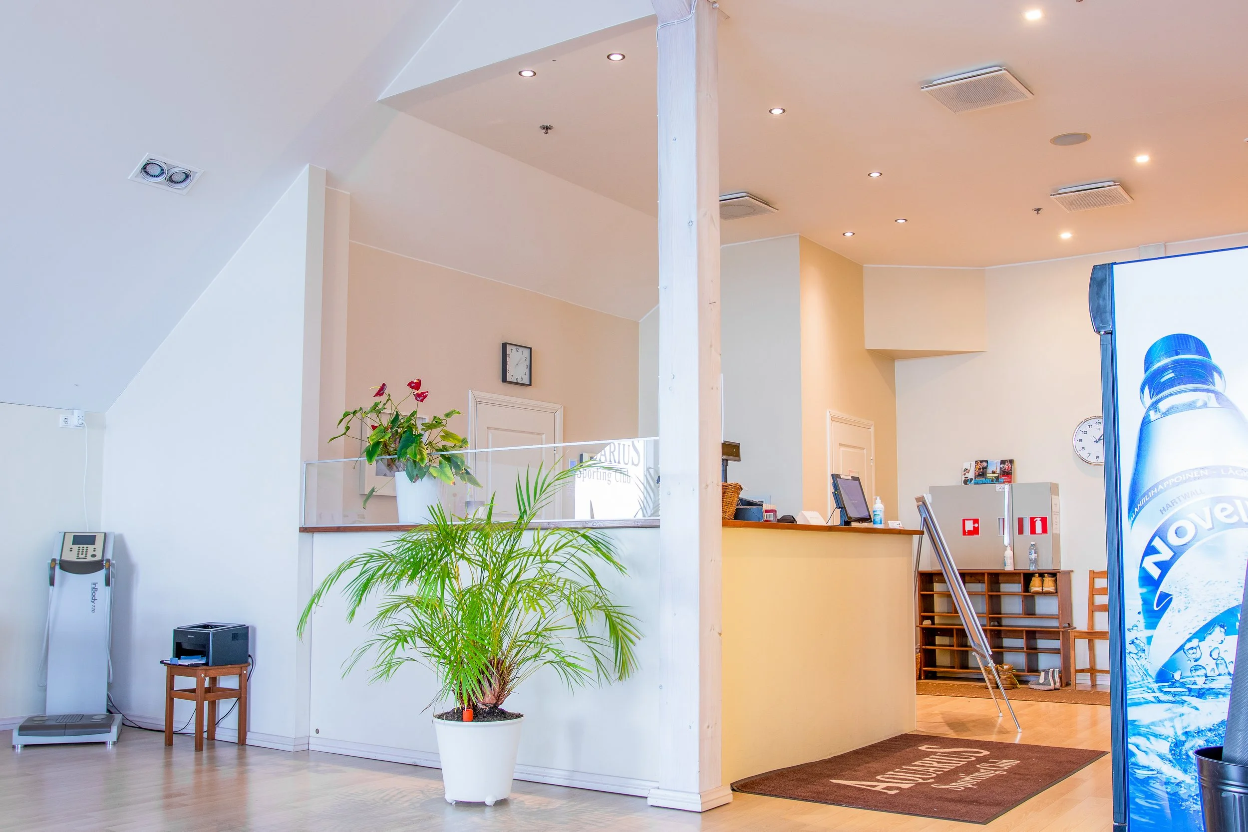 Reception area with a counter, a cash register, and a computer, flanked by potted plants, a vending machine showing bottled beverages, and seating. The space has a high ceiling, air vents, a clock, and a brown rug.