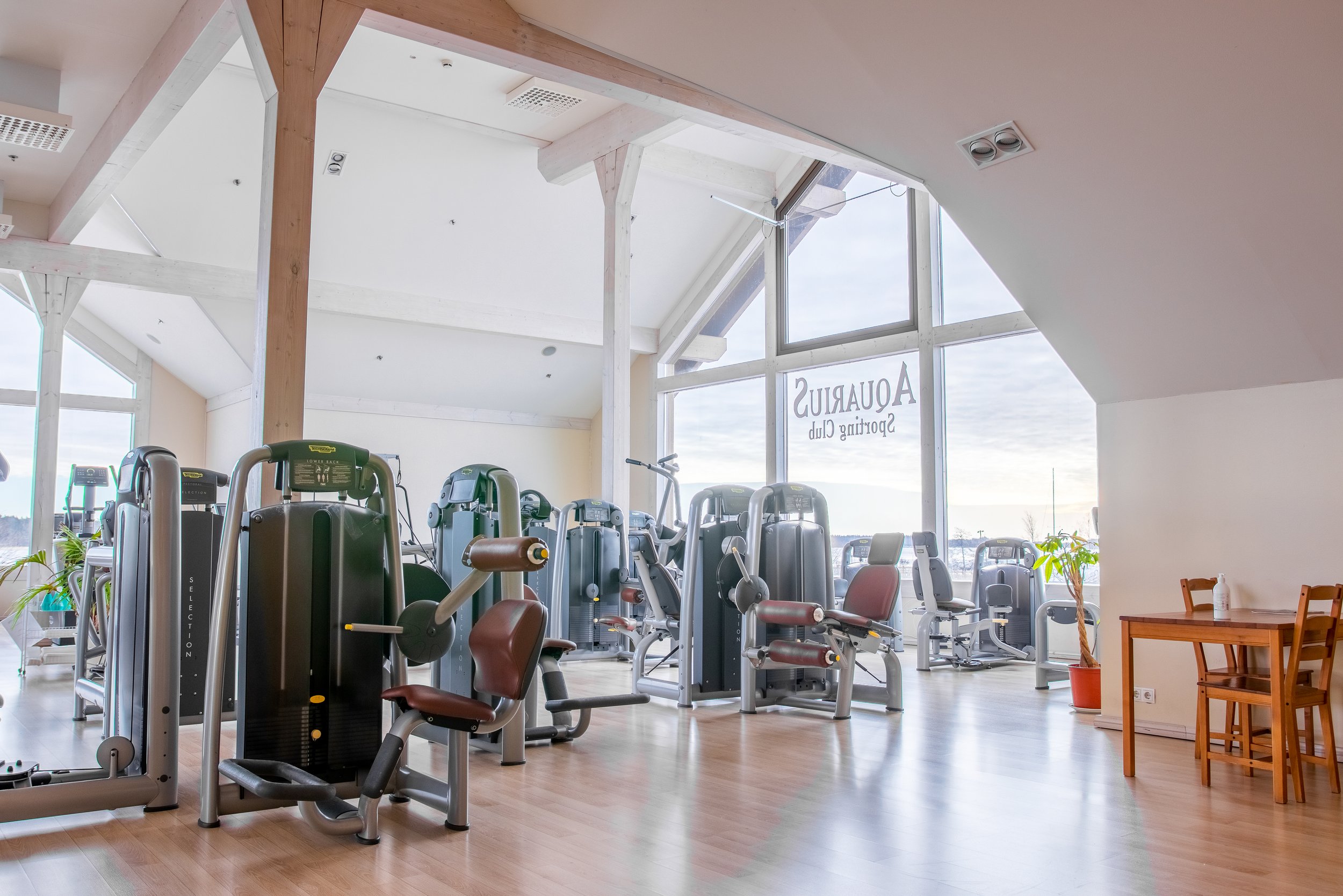 Empty gym with various exercise machines, large windows, wood flooring, and a small table with chairs in the corner.