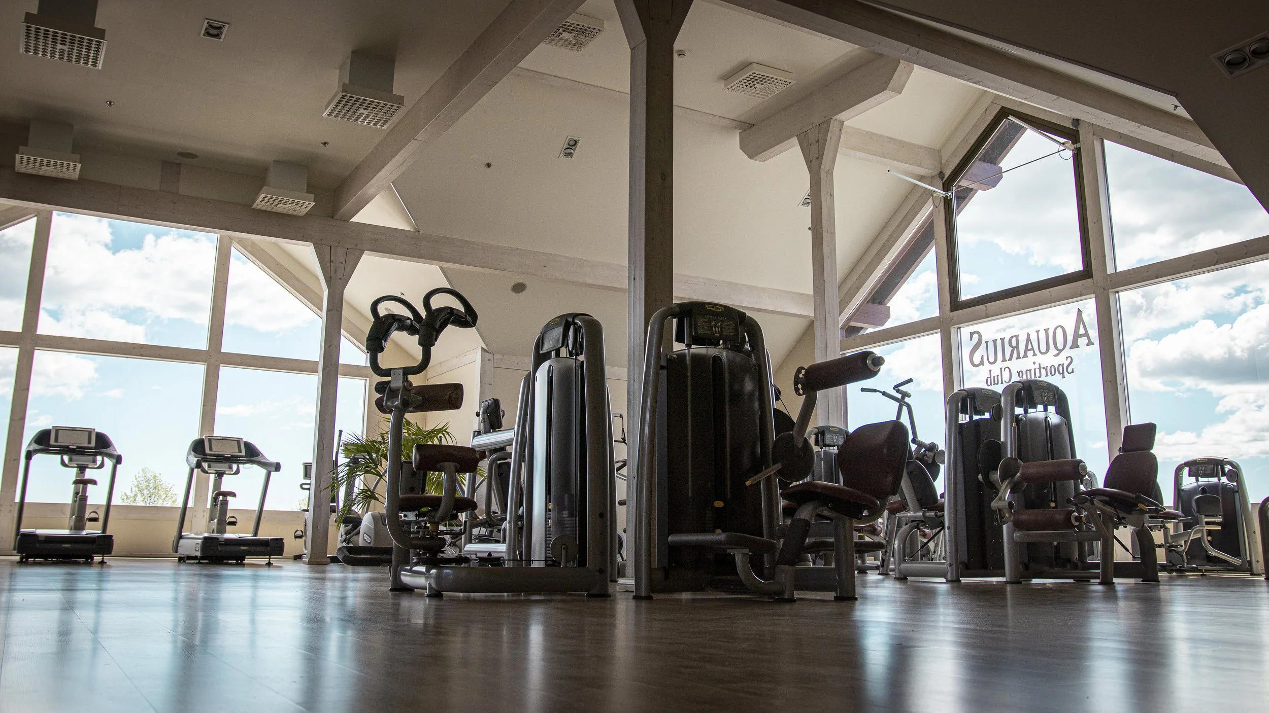 Empty gym with various exercise machines and equipment near large windows showing a blue sky with scattered clouds.