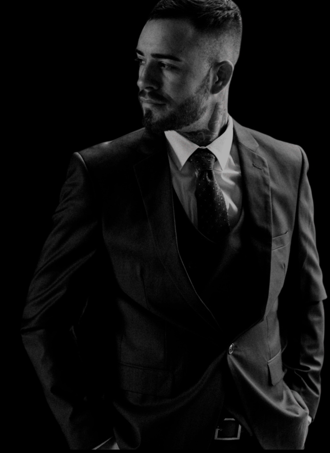 A man in a dark business suit with a tie, looking to the side, against a black background.
