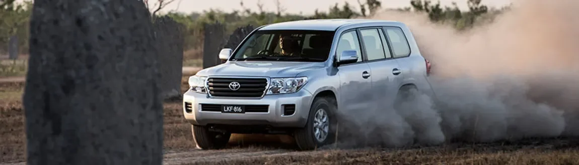 A silver Toyota LC200 driving on a dirt road kicking up dust, partially obscured by a large tree trunk in the foreground.