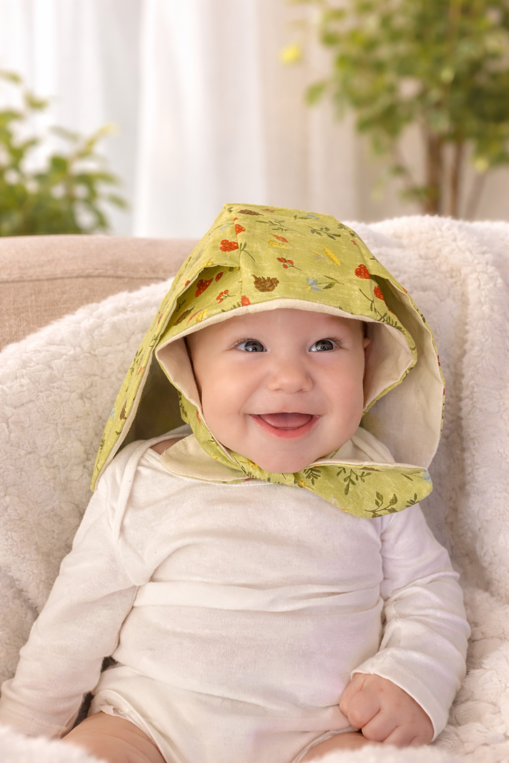 A smiling baby wearing a green bonnet with colorful floral patterns, sitting on a cozy chair with a soft blanket, in a bright indoor setting with plants in the background.