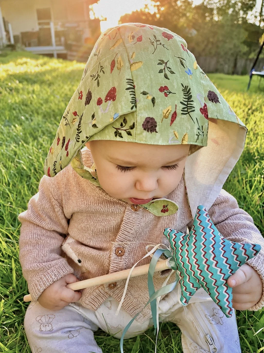 A young child sitting on the grass during sunset, wearing a green floral hat, a beige jacket, and patterned pants, holding a colorful fabric star-shaped toy with ribbons and a wooden stick.
