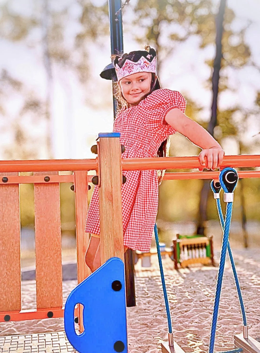A young girl wearing a red and white checkered dress and a princess crown is standing on a playground structure, holding onto a horizontal orange bar with one hand, with a background of trees and cloudy sky.