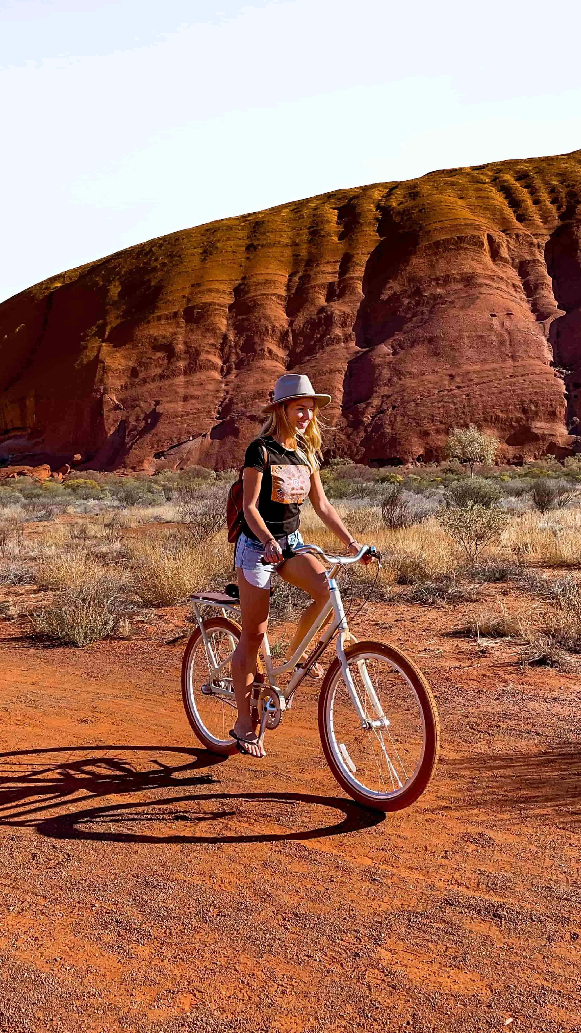 Solo podróżniczka jeździ na rowerze przy Uluru. Ilustracja wspomaga sekcję bloga podróżniczego z najciekawszymi miejscami do zobaczenia w Australii