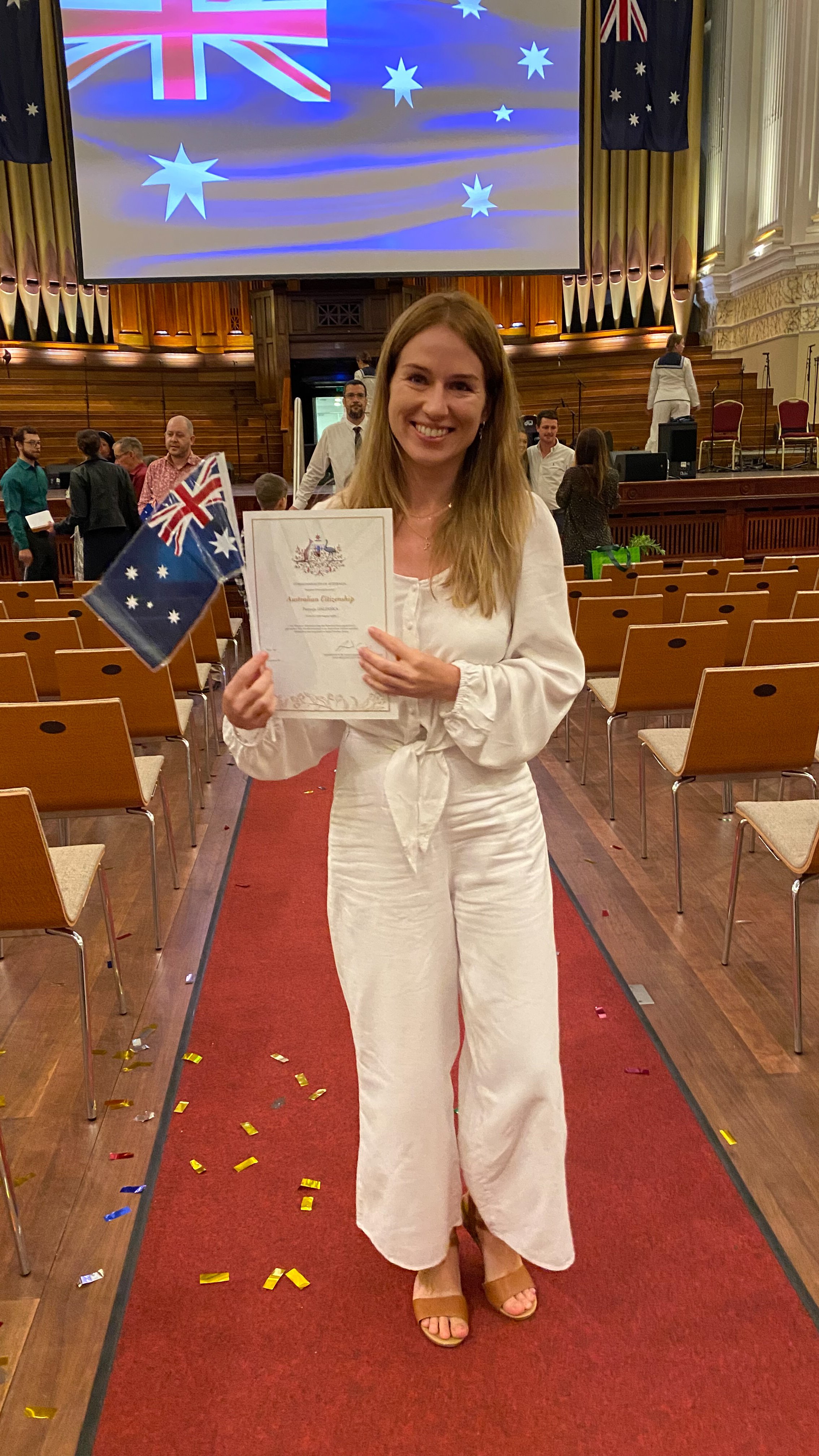 Patrycja Galinska podczas ceremonii wręczania obywatelstwa w Australii, w jednej ręce trzyma dokument potwierdzający obywatelstwo, w drugiej ręce australijską flagę. Ilustracja wspomaga opis jak dostać obywatelstwo w Australii