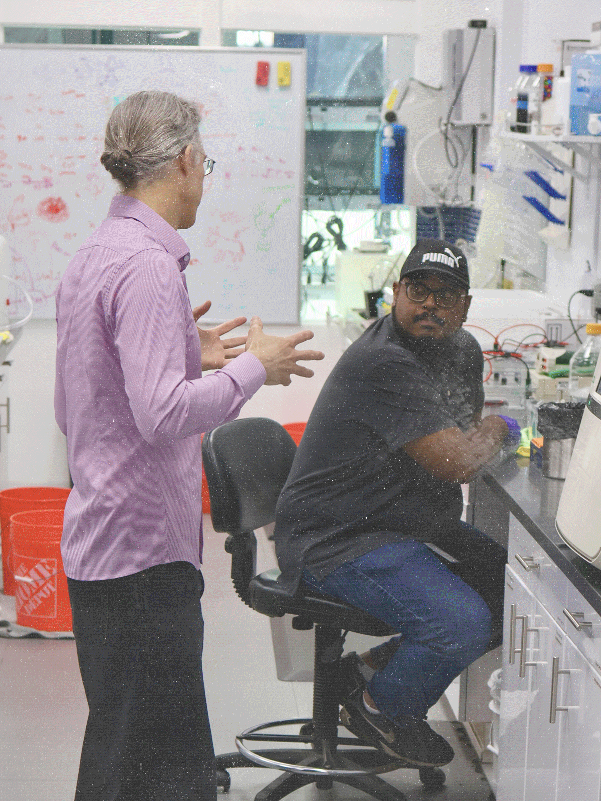 Two scientists work in a laboratory, one standing and the other sitting at a lab bench, with scientific equipment and a whiteboard in the background.