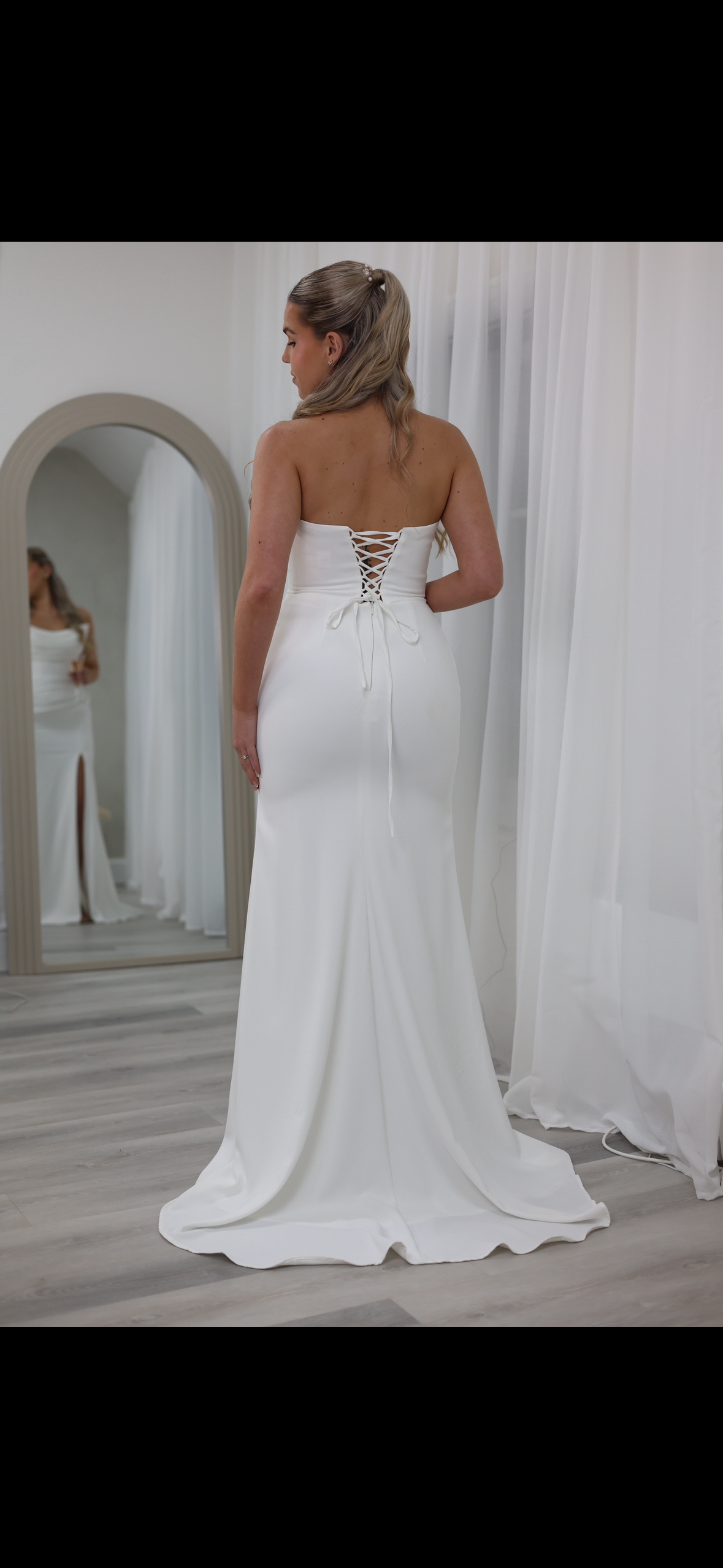 A woman in a white wedding dress with a lace-up back is standing in front of a mirror, adjusting her dress.