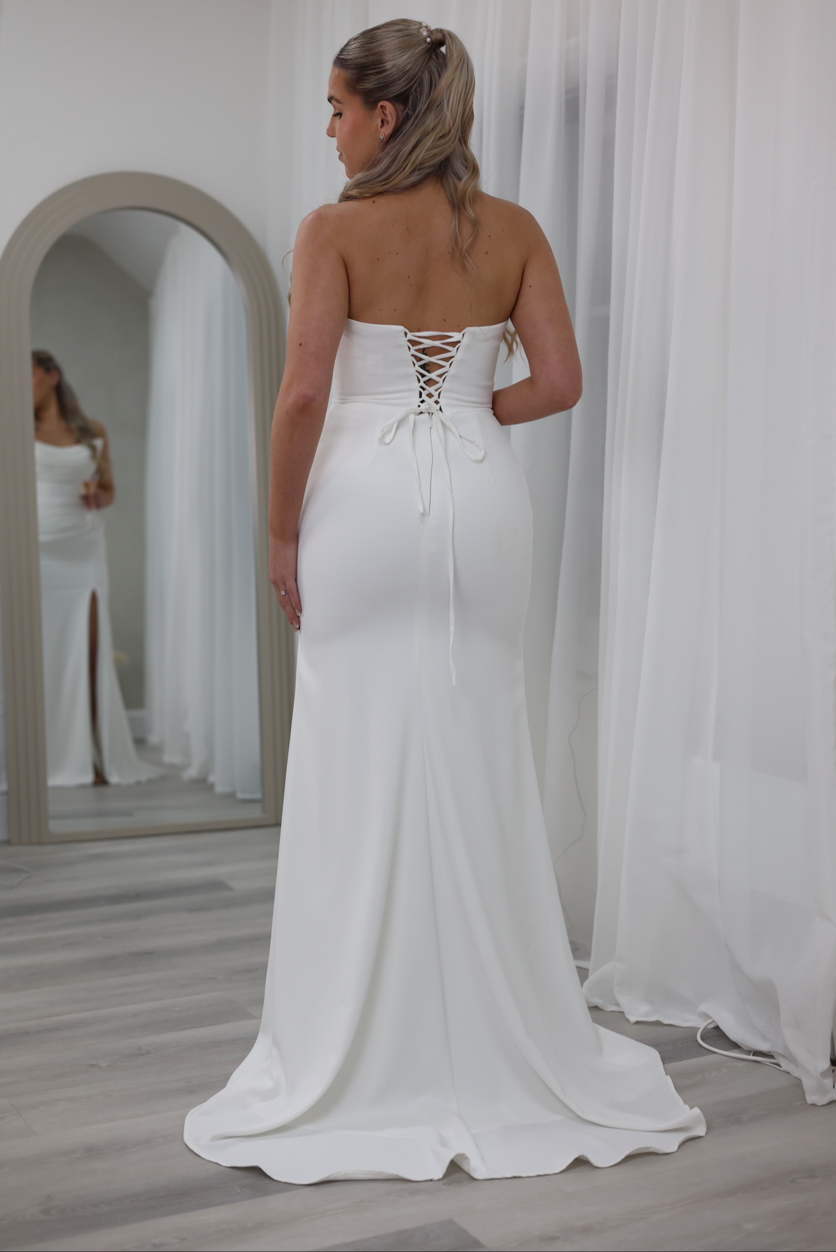 A woman trying on a white strapless wedding dress with a lace-up back, standing in front of a mirror in a fitting room.