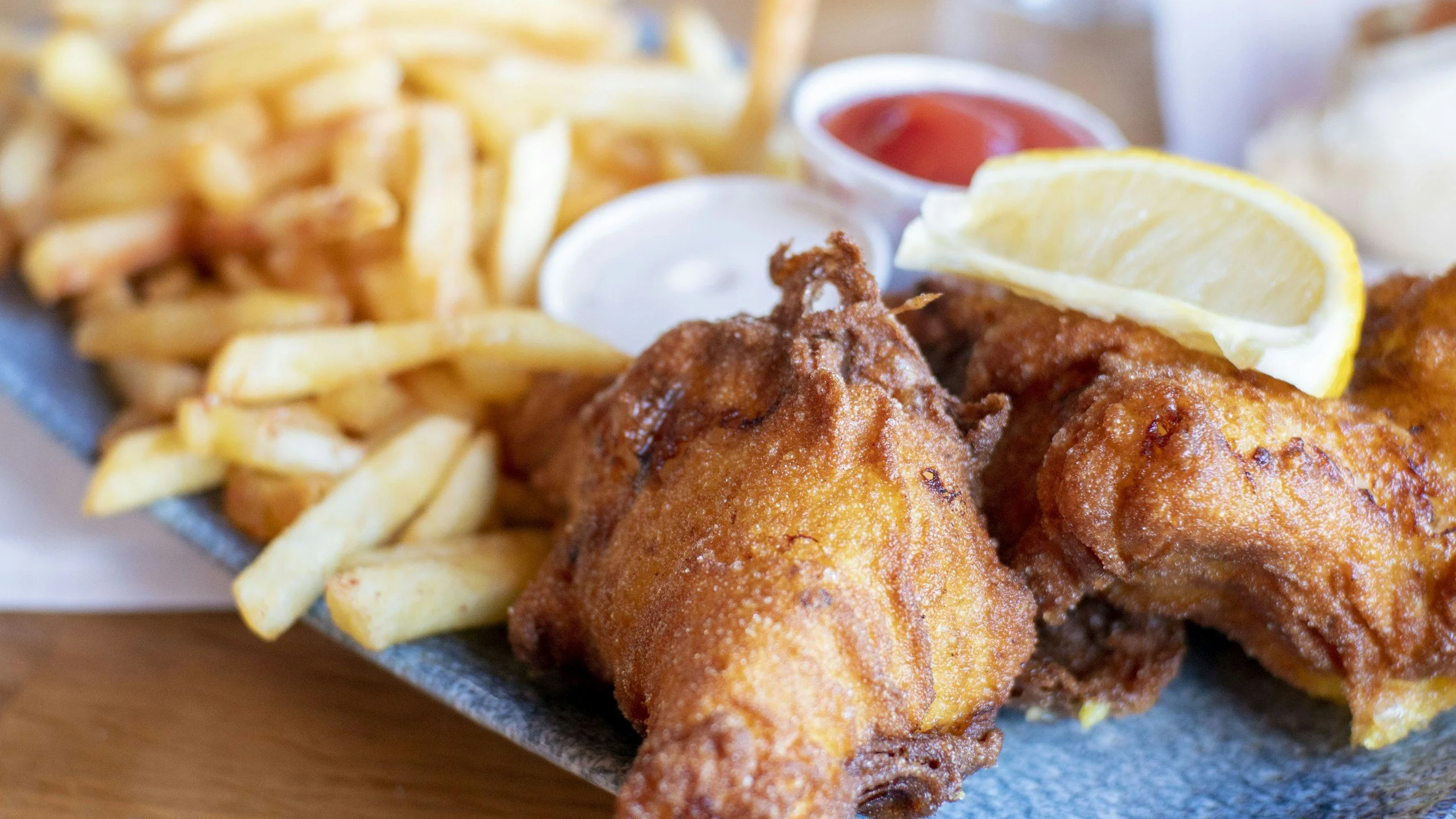 Close-up of golden battered fish and chips with a lemon wedge, ketchup, and tartar sauce on a blue plate.