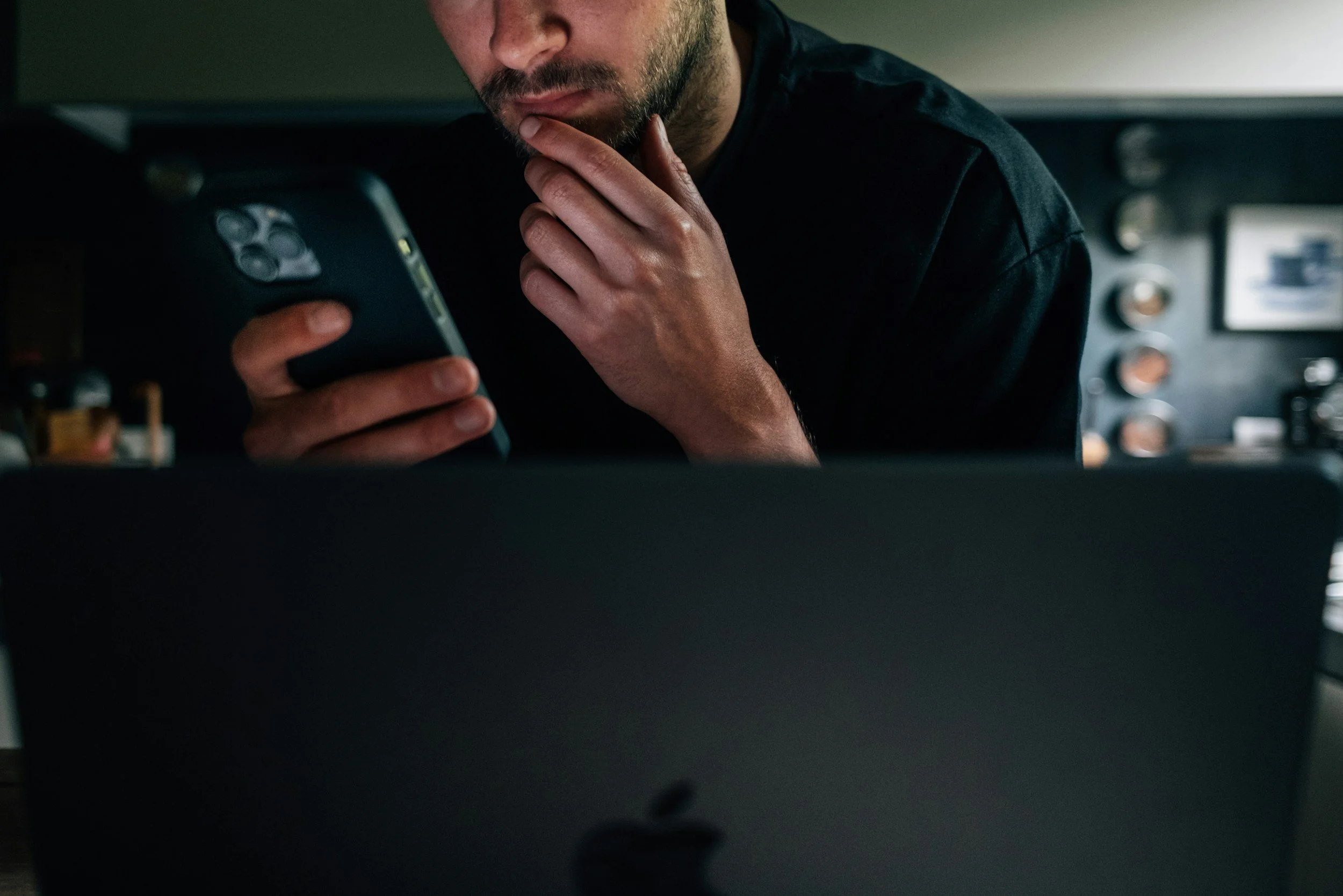 A man with a beard looking at his phone while sitting in front of an Apple MacBook.