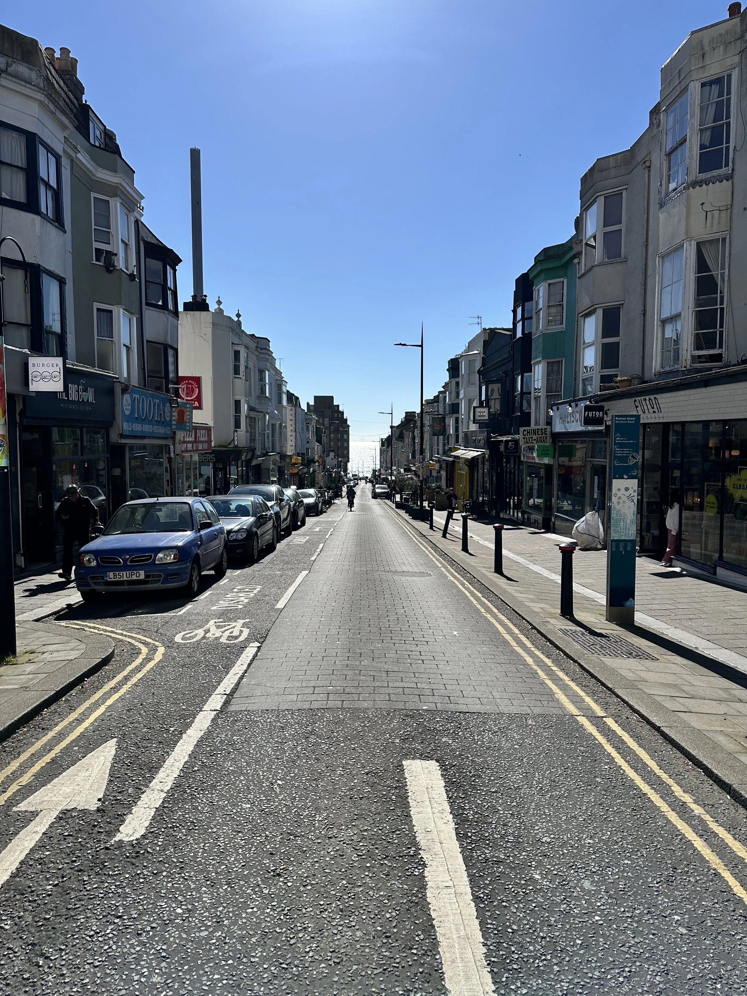 View down Preston Street in Brighton toward the sea with independent shops, parked cars, and a cyclist.