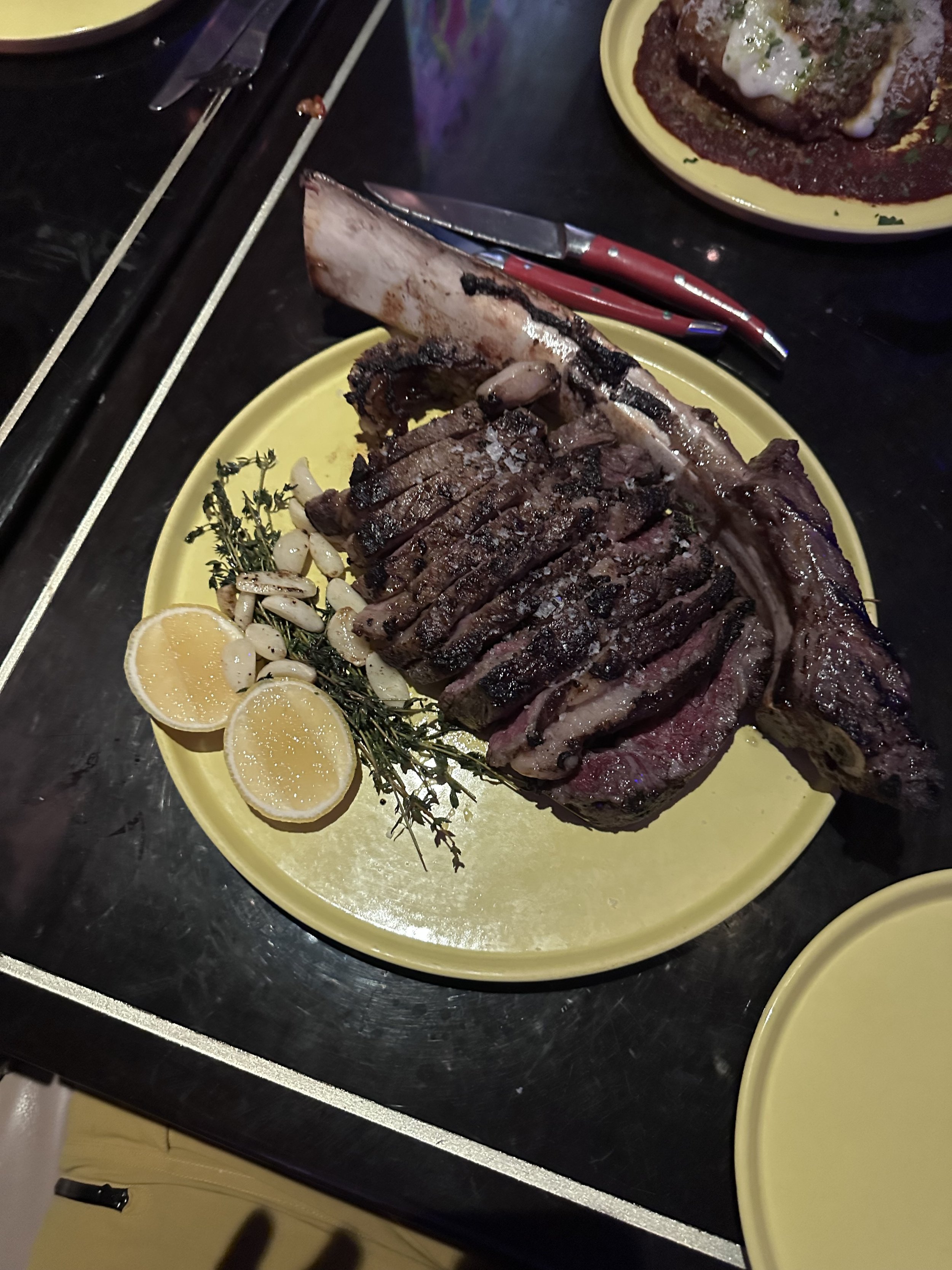 Sliced tomahawk ribeye steak served on a yellow plate with roasted garlic, herbs, and lemon at Superfrico Las Vegas.