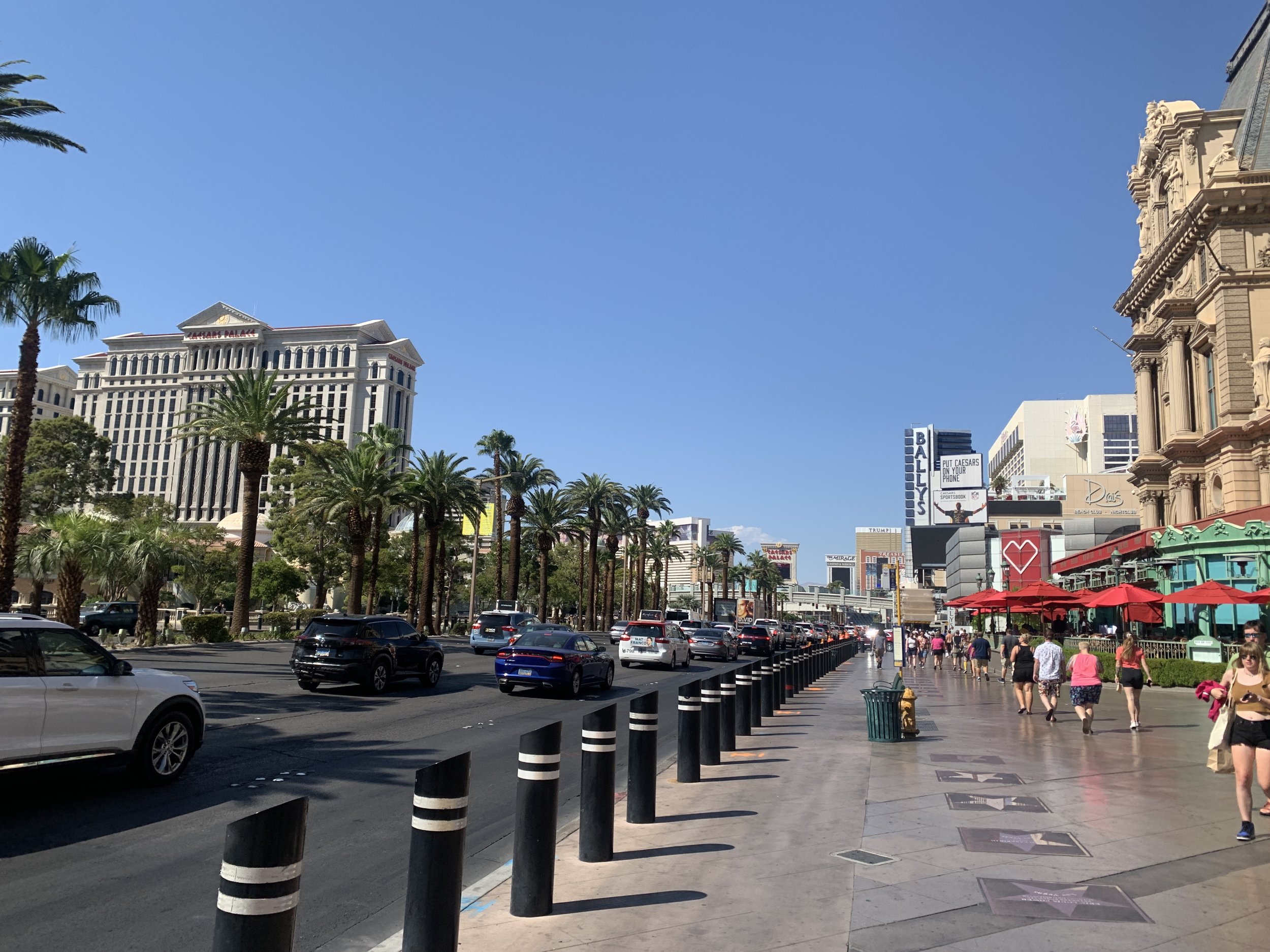 Traffic on the Las Vegas Strip next to a crowded pedestrian sidewalk with palm trees and red restaurant umbrellas.