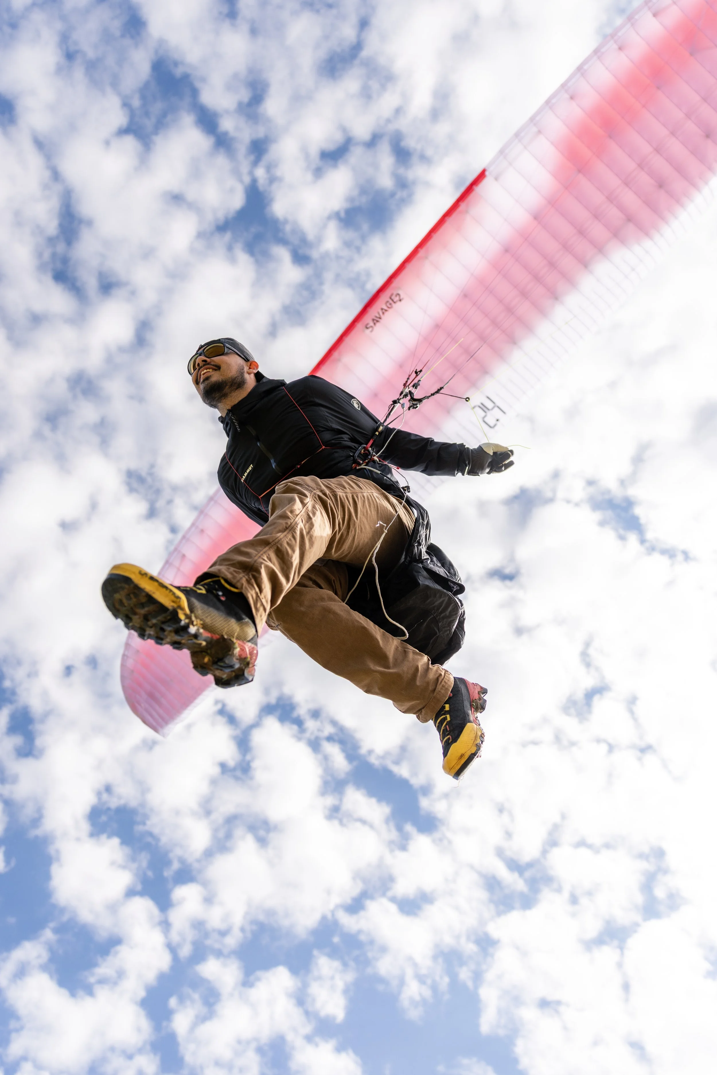 A person is paragliding through a sky with white clouds, wearing sunglasses, a black jacket, and brown pants.