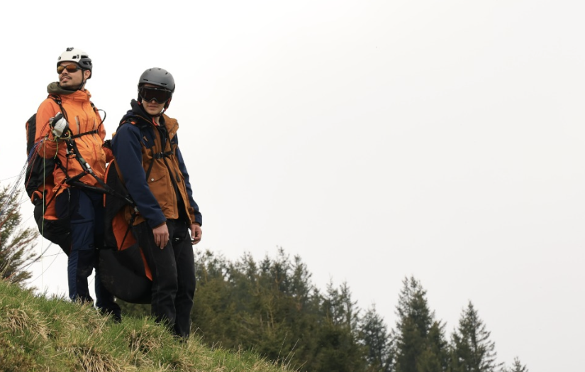 Two paragliders standing on a grassy hill with a forested landscape in the background, wearing helmets and outdoor gear.