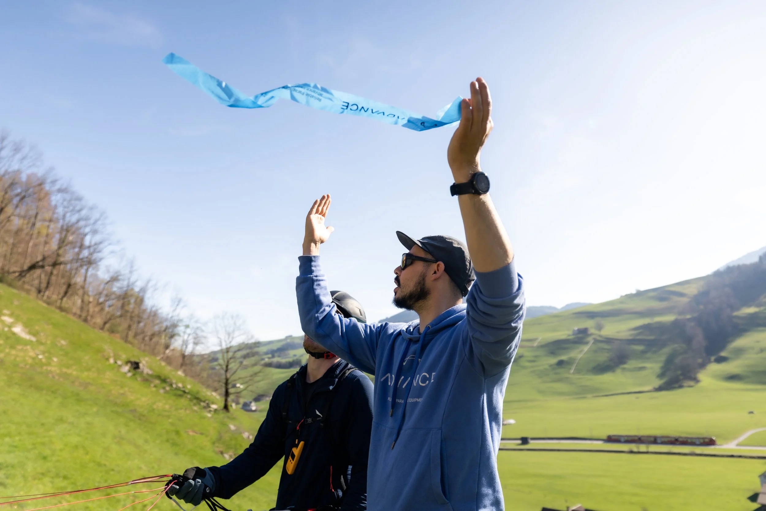 A man wearing sunglasses and a cap flying a kite outdoors in a grassy area with rolling hills and trees in the background.