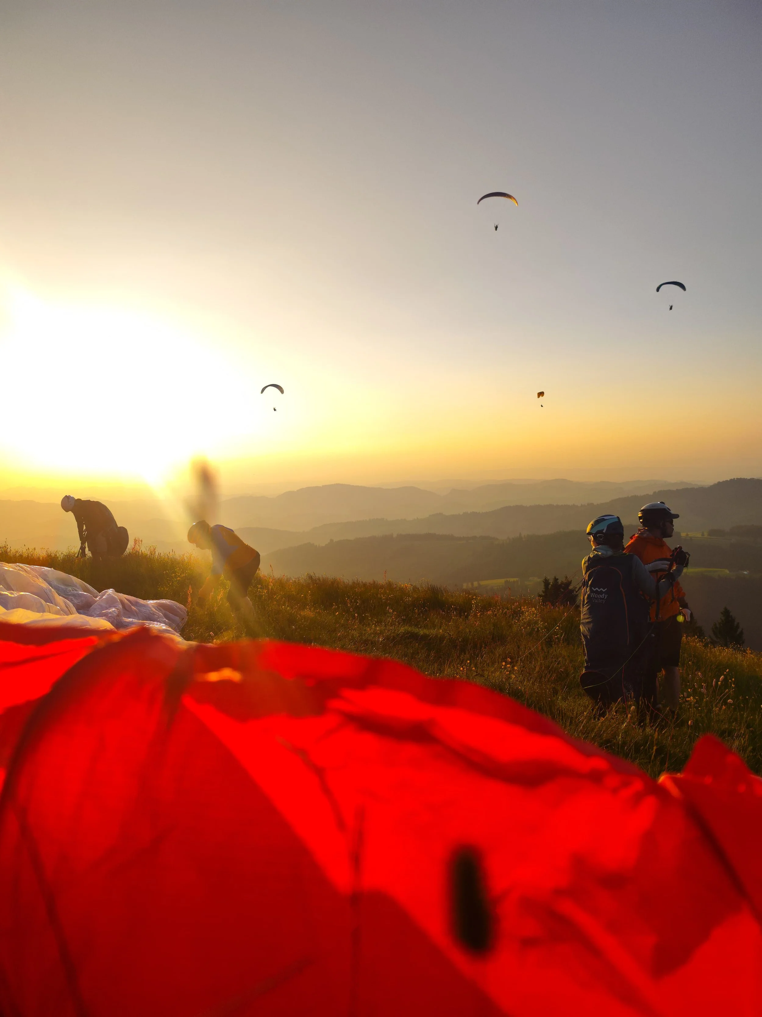 A group of people preparing for a parachute jump during sunset in a scenic outdoor landscape with hills and scattered trees, with some individuals wearing helmets and gear and parachutes laid out on the ground.