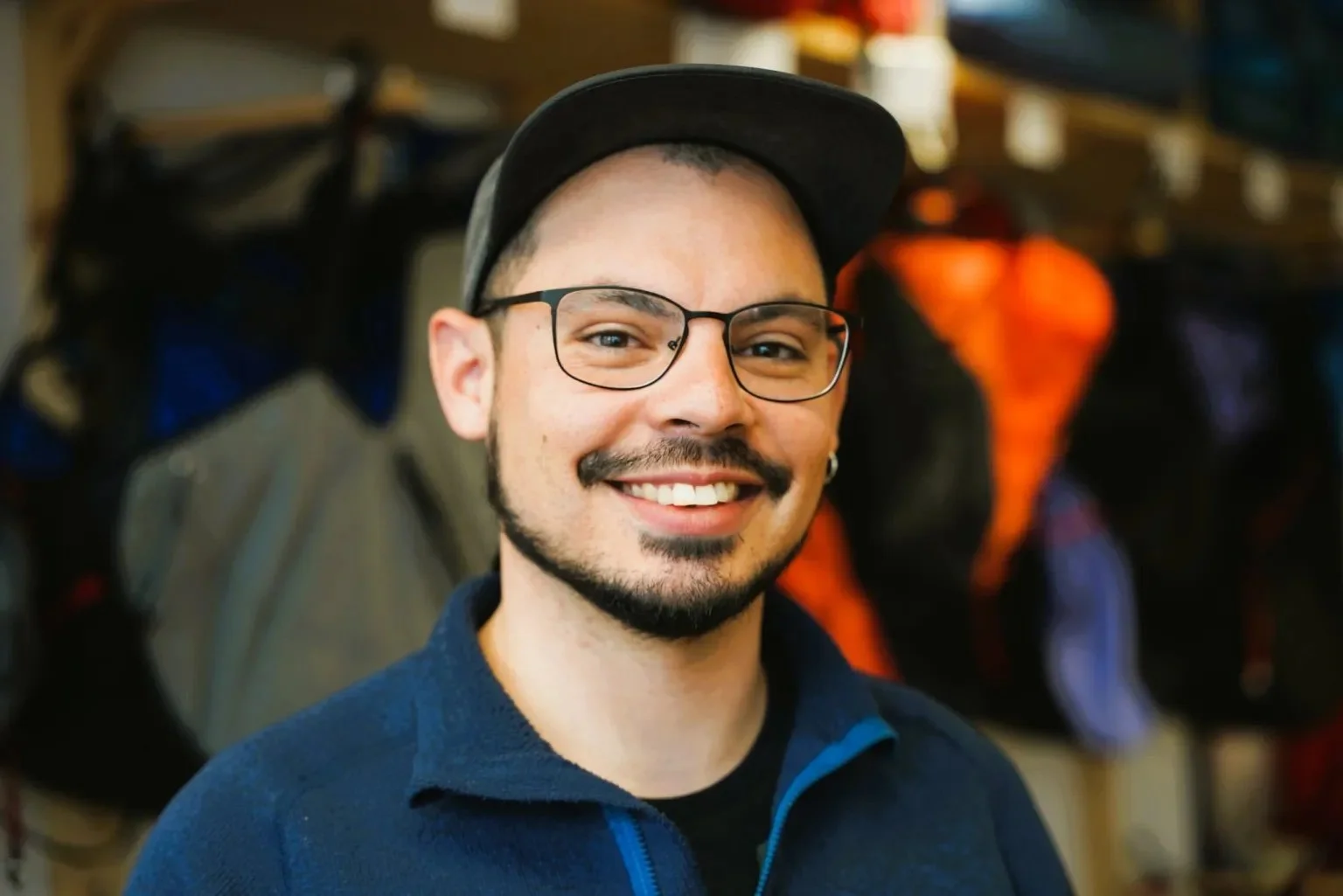Smiling man with glasses and a beard, wearing a black cap and a blue jacket, indoors with backpacks hanging on the wall behind him.
