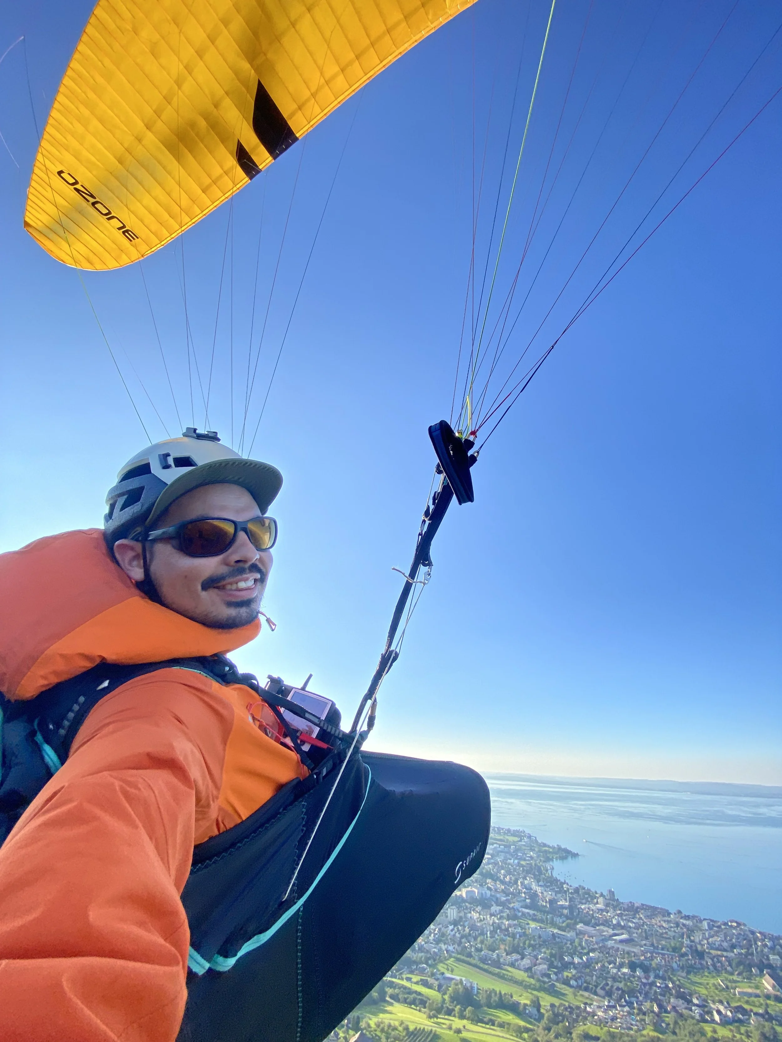 A person in an orange jacket, wearing sunglasses and a helmet, appears to be paragliding above a city landscape on a clear day with blue sky and water in the background.