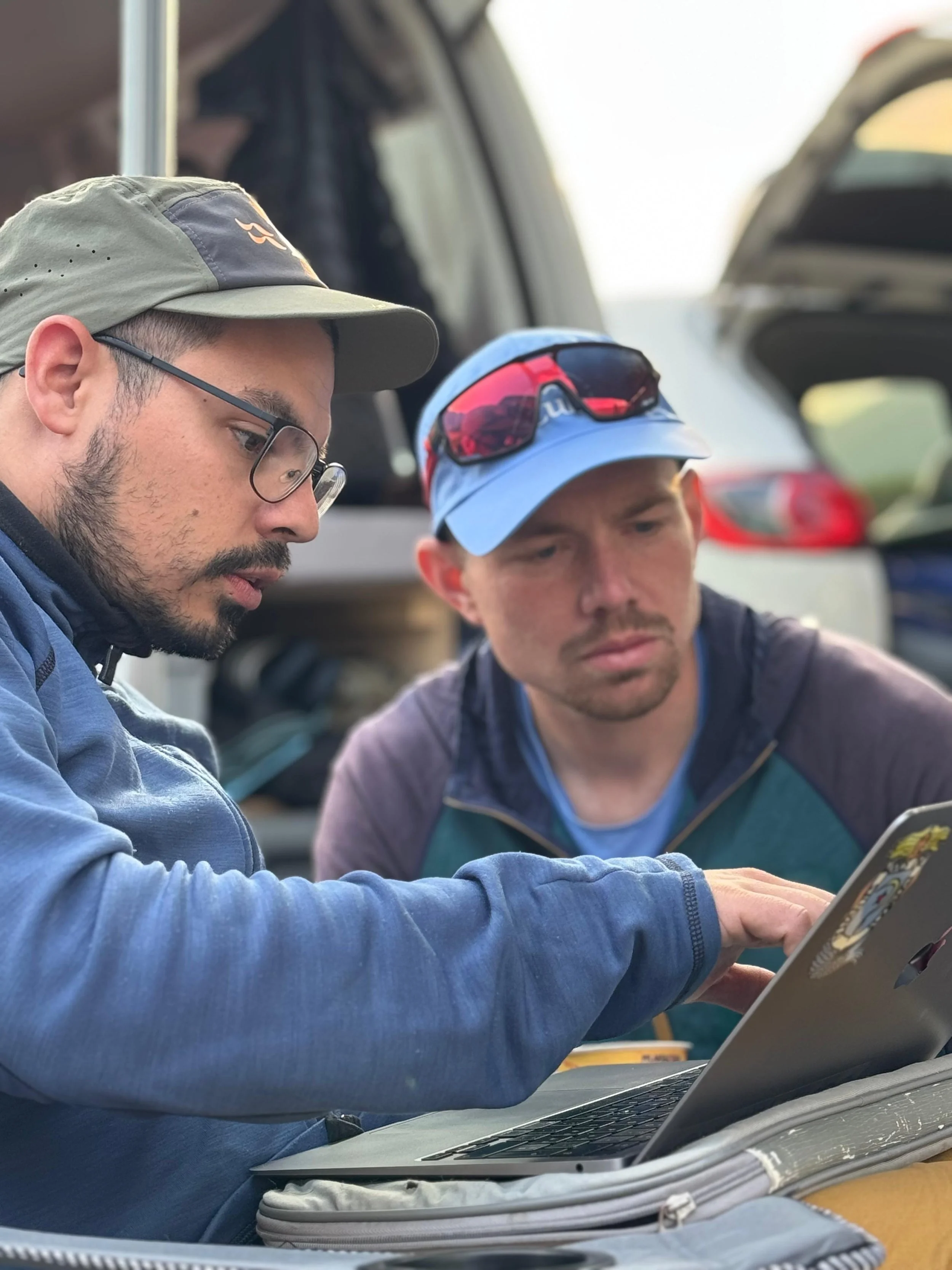 Two men sitting closely together outdoors, looking at a tablet device, with an open car trunk in the background.