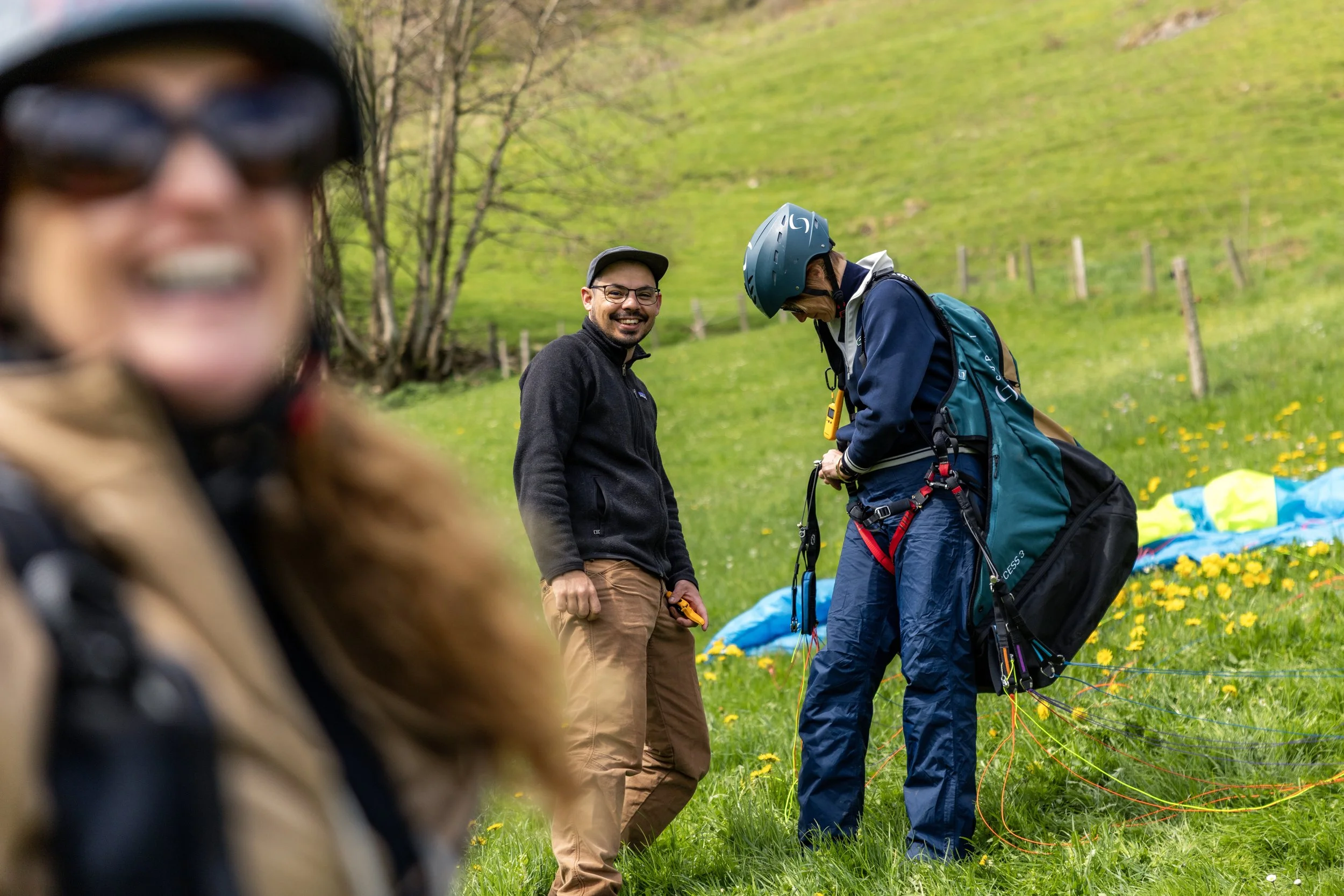 Three people outdoors on a grassy hillside with trees in the background. One person in the foreground is blurred and wearing sunglasses and a helmet. The other two are in the middle distance, smiling; one is wearing a baseball cap and glasses, and the other is wearing a helmet and harness, preparing for outdoor activity.