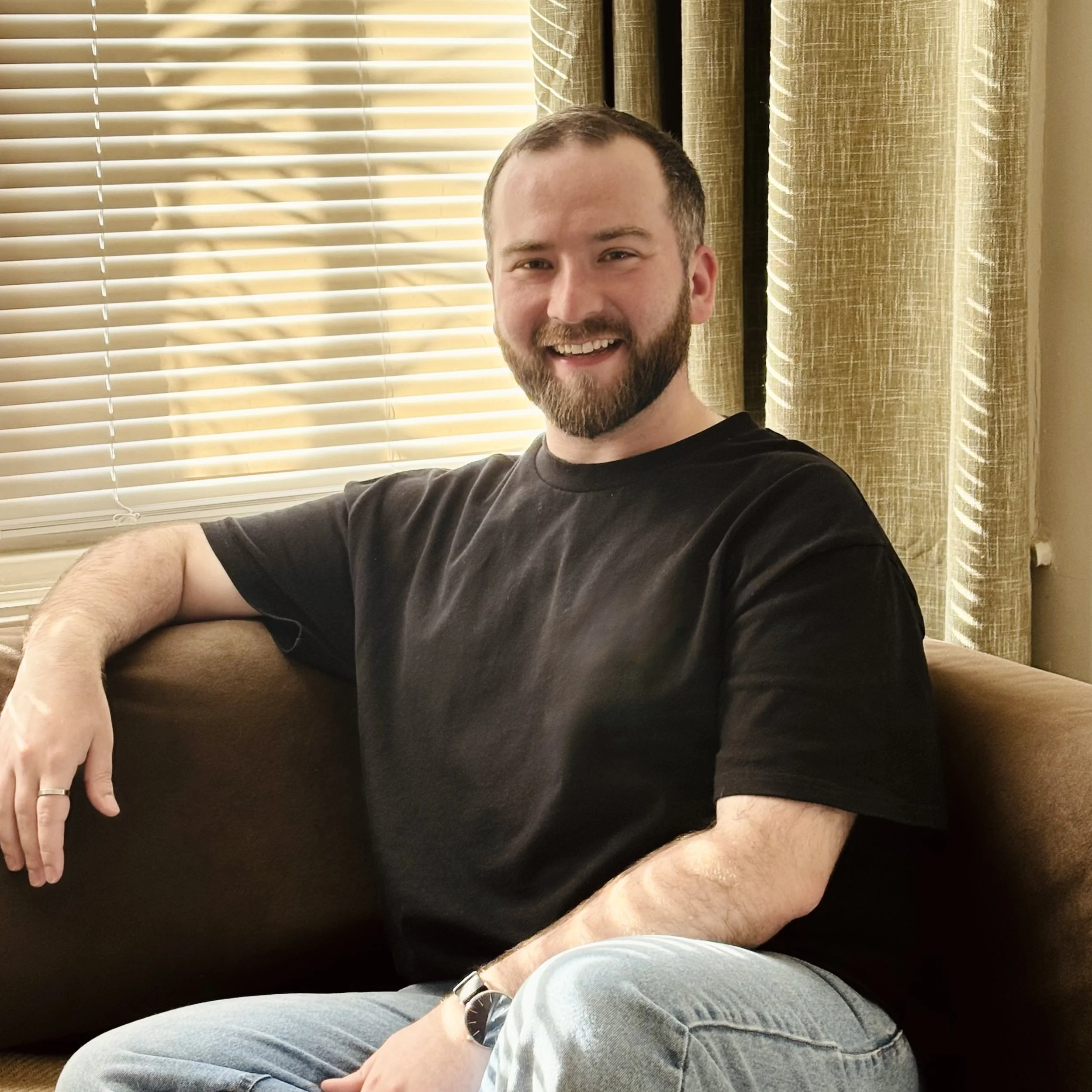 A smiling man with a beard wearing a black t-shirt, sitting on a brown couch in front of closed window blinds and beige curtains.