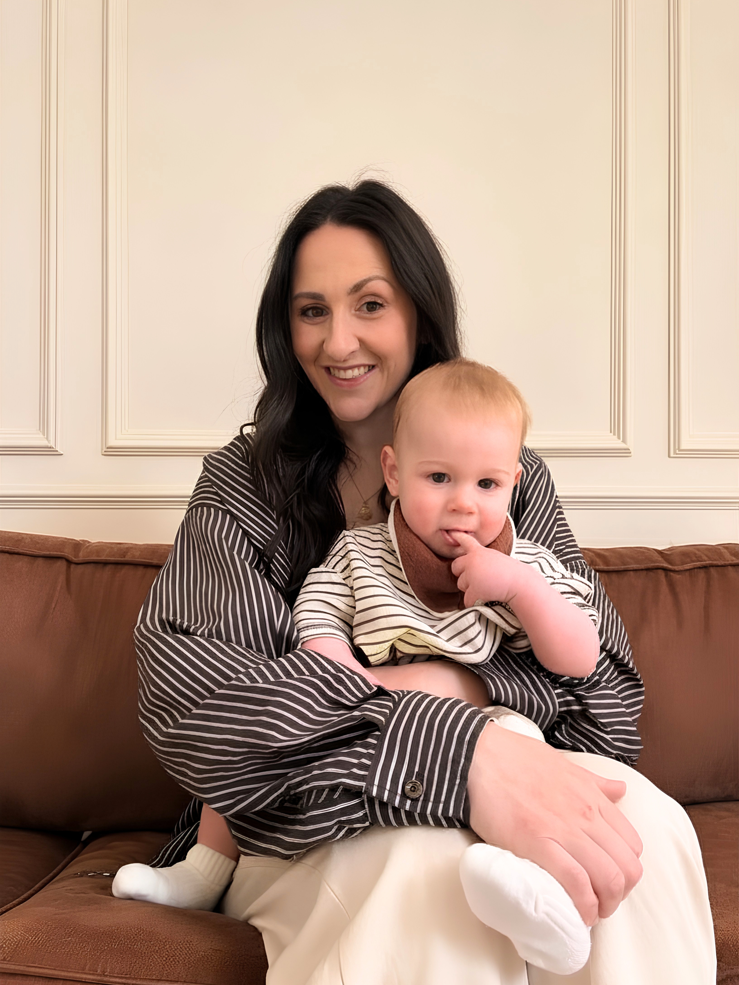 A woman with long dark hair smiling while sitting on a brown couch, holding a child with light hair and fair skin, who is touching his mouth with his finger, with a white wall with decorative molding in the background.