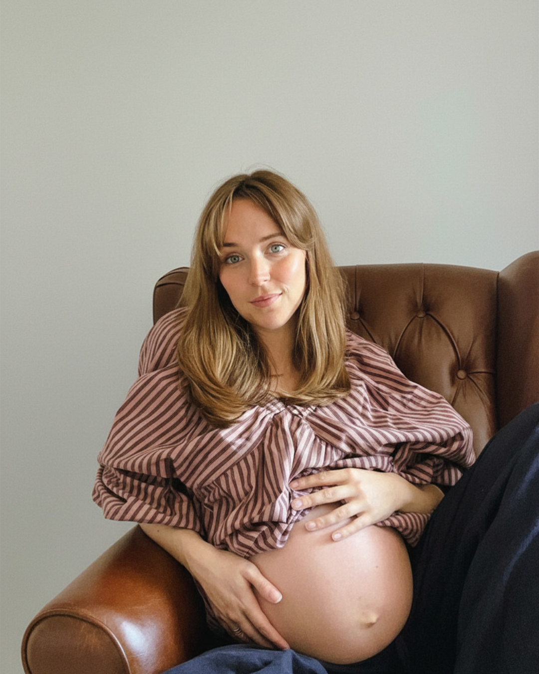 A pregnant woman with long light brown hair sitting on a brown leather couch, wearing a pink and brown striped top, resting her hand on her belly and looking at the camera with a slight smile.
