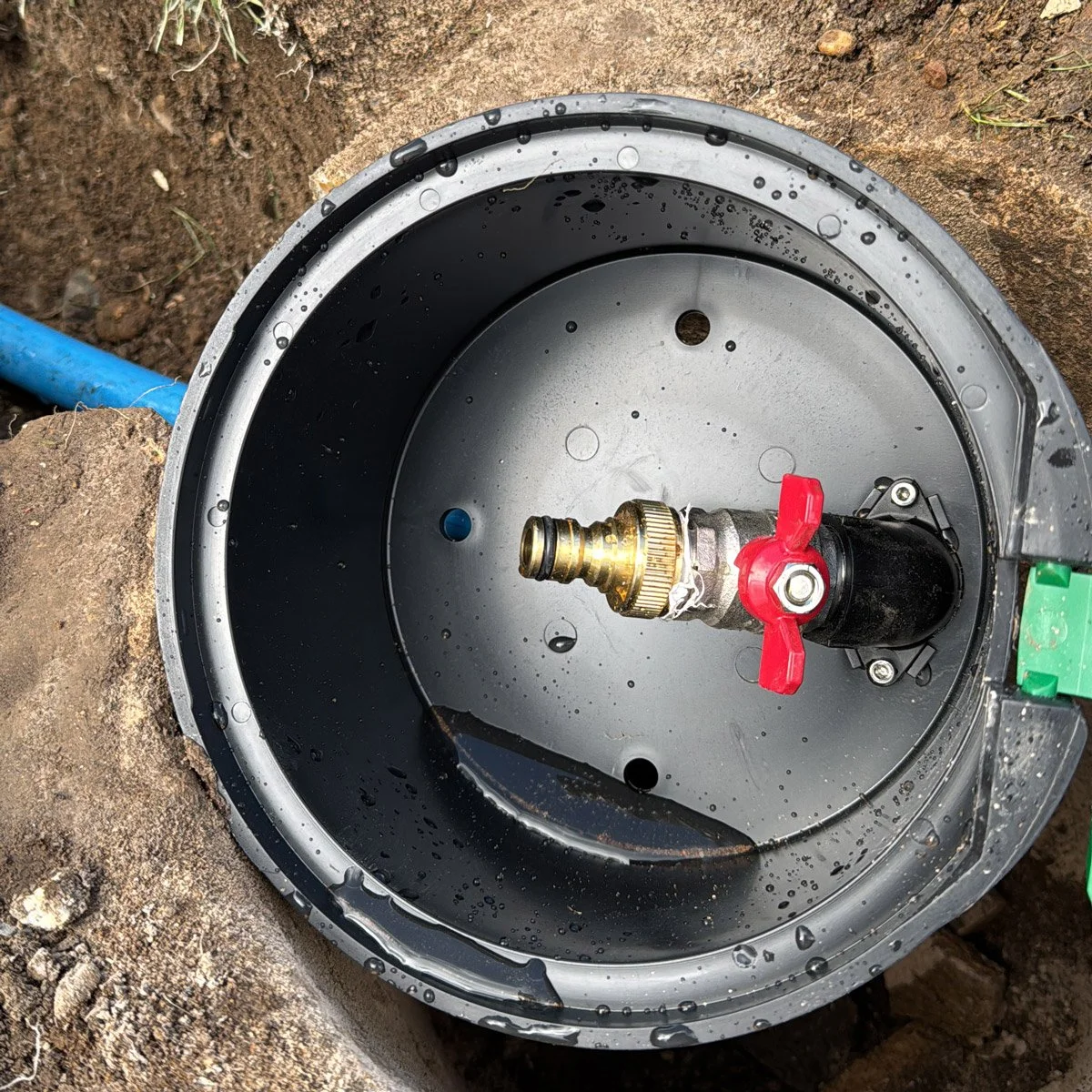 A black and silver irrigation timer with a brass connector and red valve handle installed inside a plastic housing, with water droplets on the surface, positioned on soil near blue piping.
