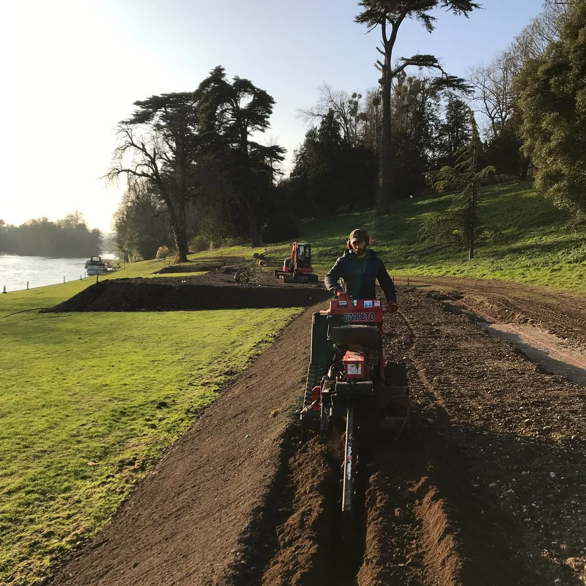 laying trenches for water pipes as part of an irrigation system near the Thames, with trees and grassy areas in the background.