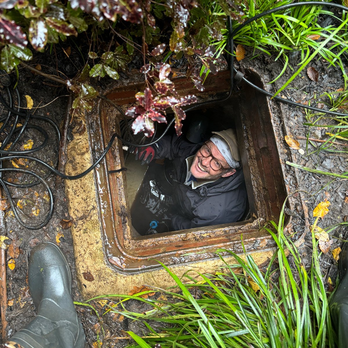 Person smiling while fitting an irrigation system standing in a water tank, surrounded by muddy ground and green plants, wearing glasses, a bandana, waterproof jacket, and gloves.
