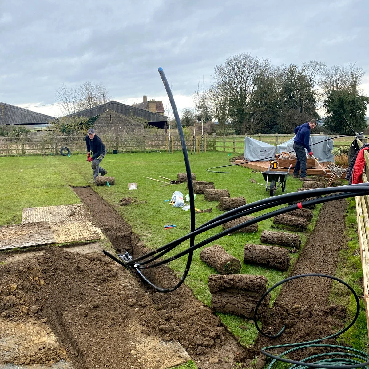 Two men are working on an irrigation project, digging trenches for a specialist bespoke water system. Tools and construction materials are scattered around, with a wheelbarrow and covered wooden deck in the background.