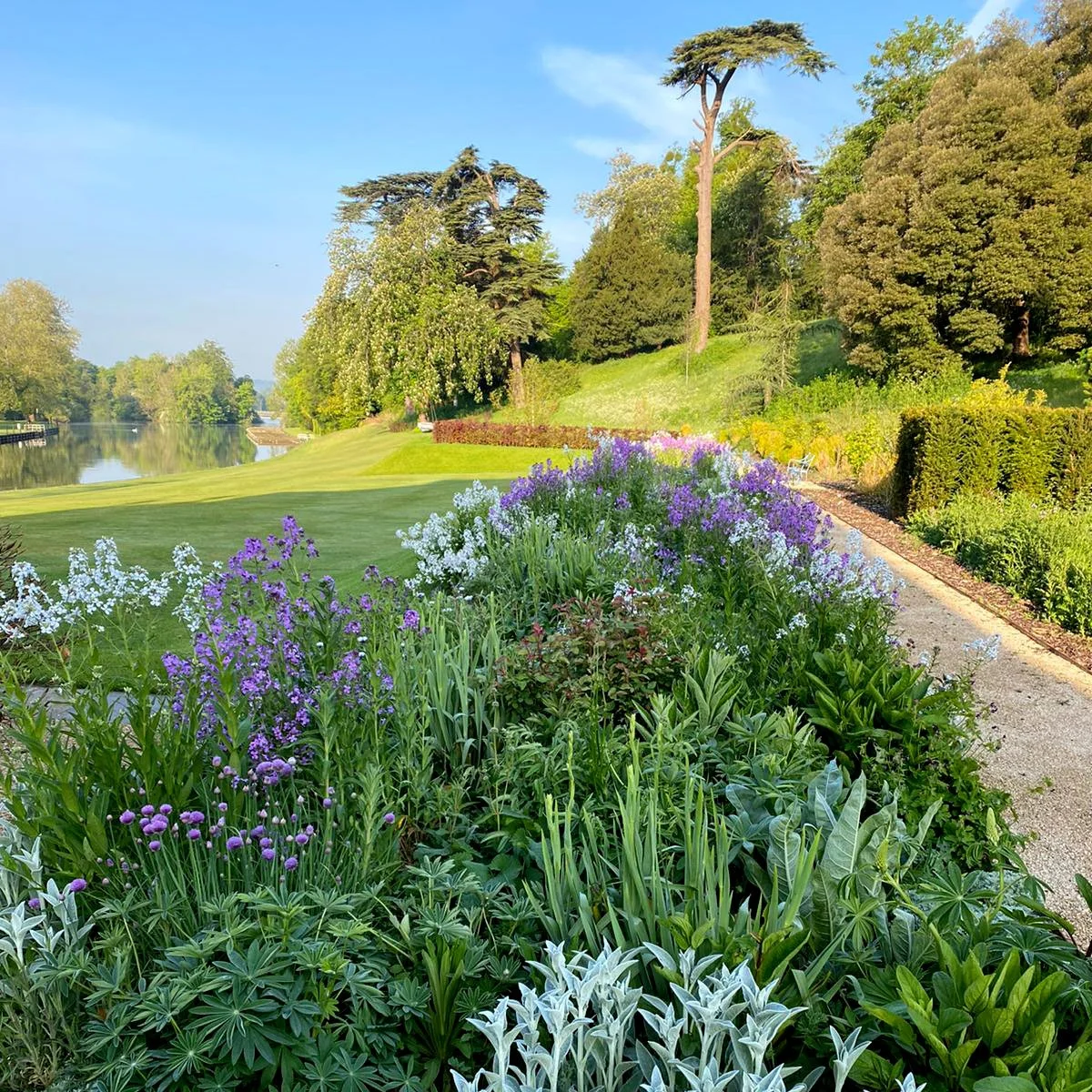 Scenic view of a garden with irrigation system by a river, with colourful flowers and lush green trees under a clear blue sky.